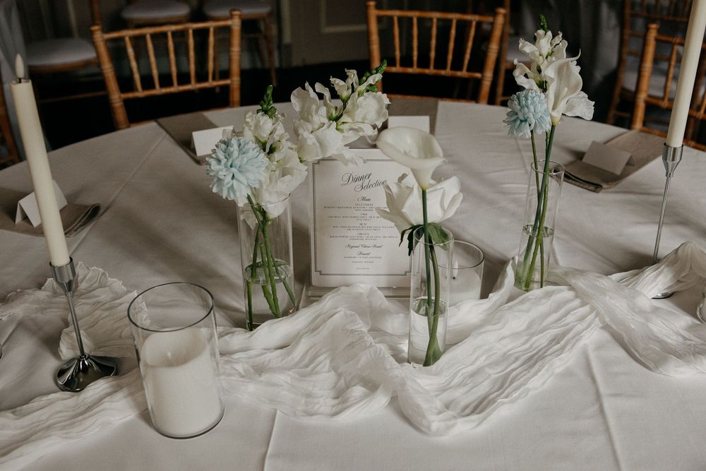 Elegant wedding table centerpiece with white flowers in bud vases, taper candles, and a menu card on white linen.