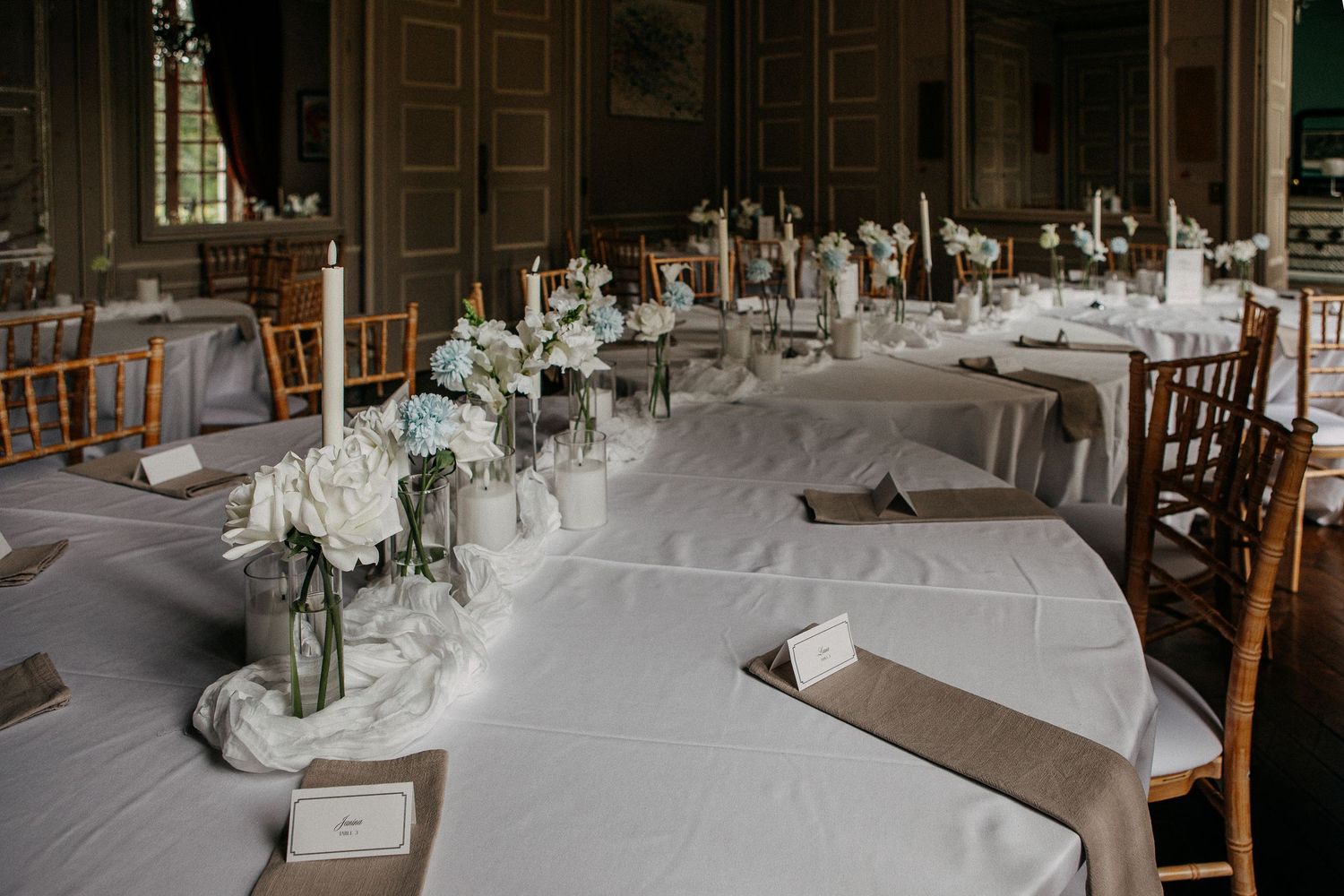 Elegant wedding reception hall with white tablecloths, floral centerpieces, and chiavari chairs in a grand venue.