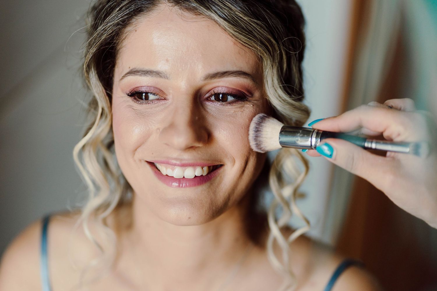 Smiling bride getting blush applied with a makeup brush during professional bridal makeup preparation.