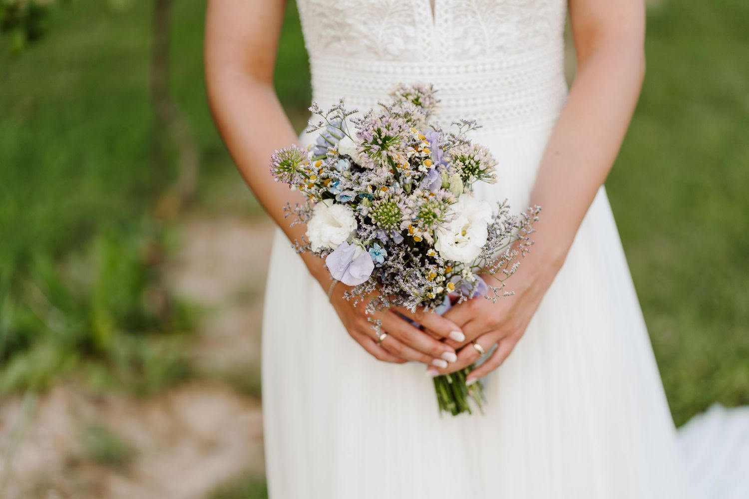 Bride in white wedding dress holding a delicate bouquet of lavender, white roses, and wildflowers outdoors.