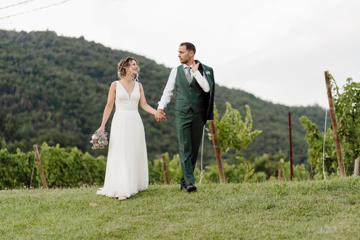Bride and groom walk hand in hand through lush vineyard with green hills backdrop on their wedding day.