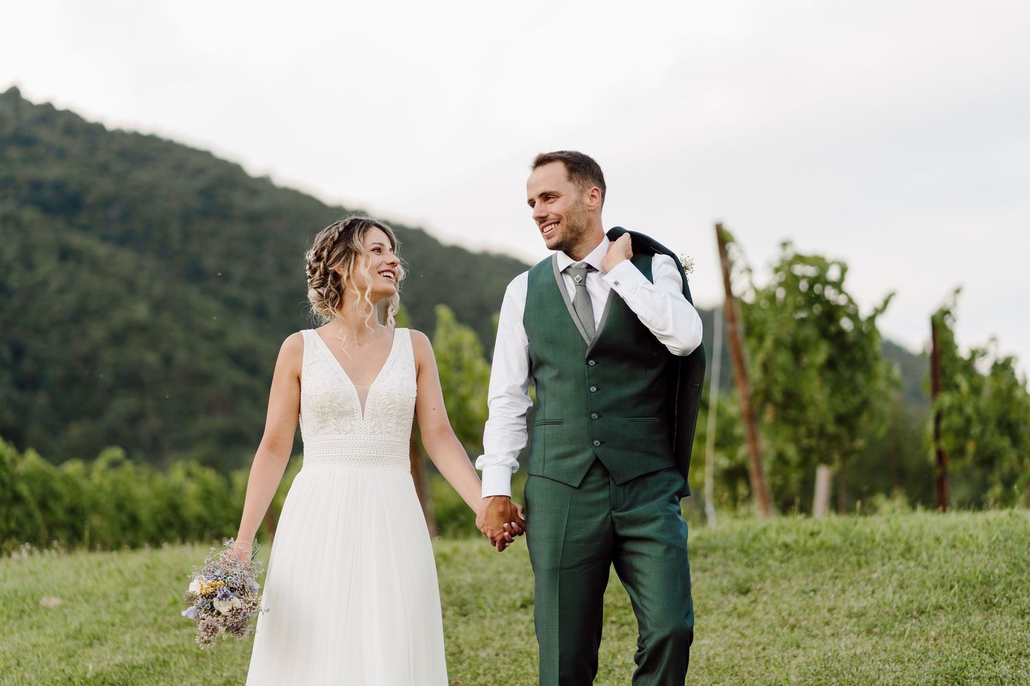 A bride and groom walk hand in hand through a lush vineyard, smiling at each other on their wedding day.