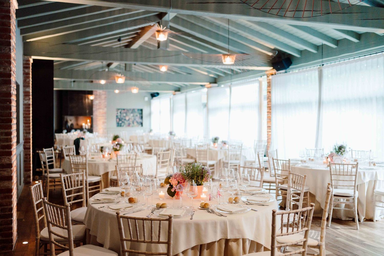 Elegant wedding reception hall with round tables, chiavari chairs, floral centerpieces, and exposed beam ceiling with pendant lights.