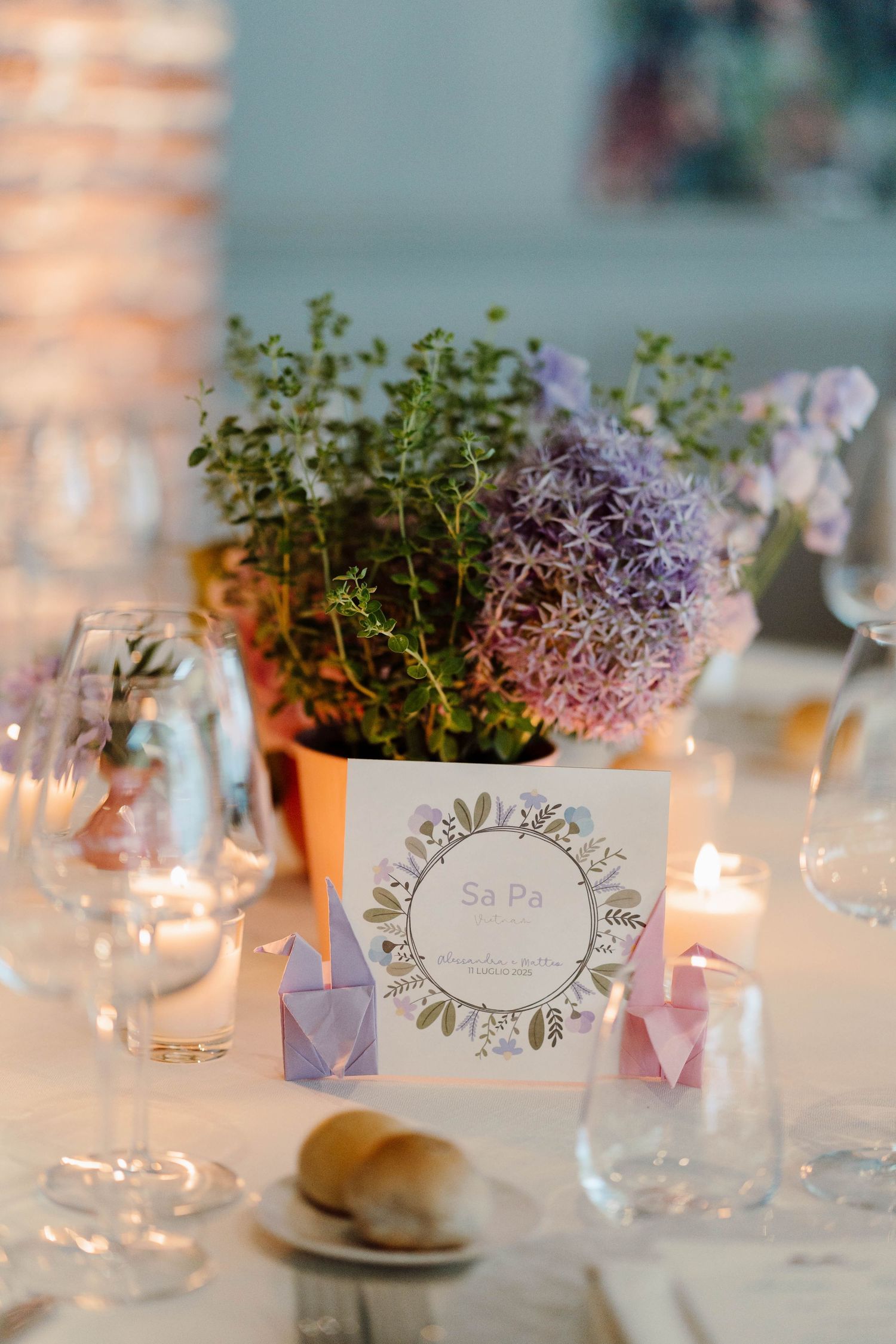Elegant wedding table setting with purple allium flowers, origami decorations, candles, and a floral place card.