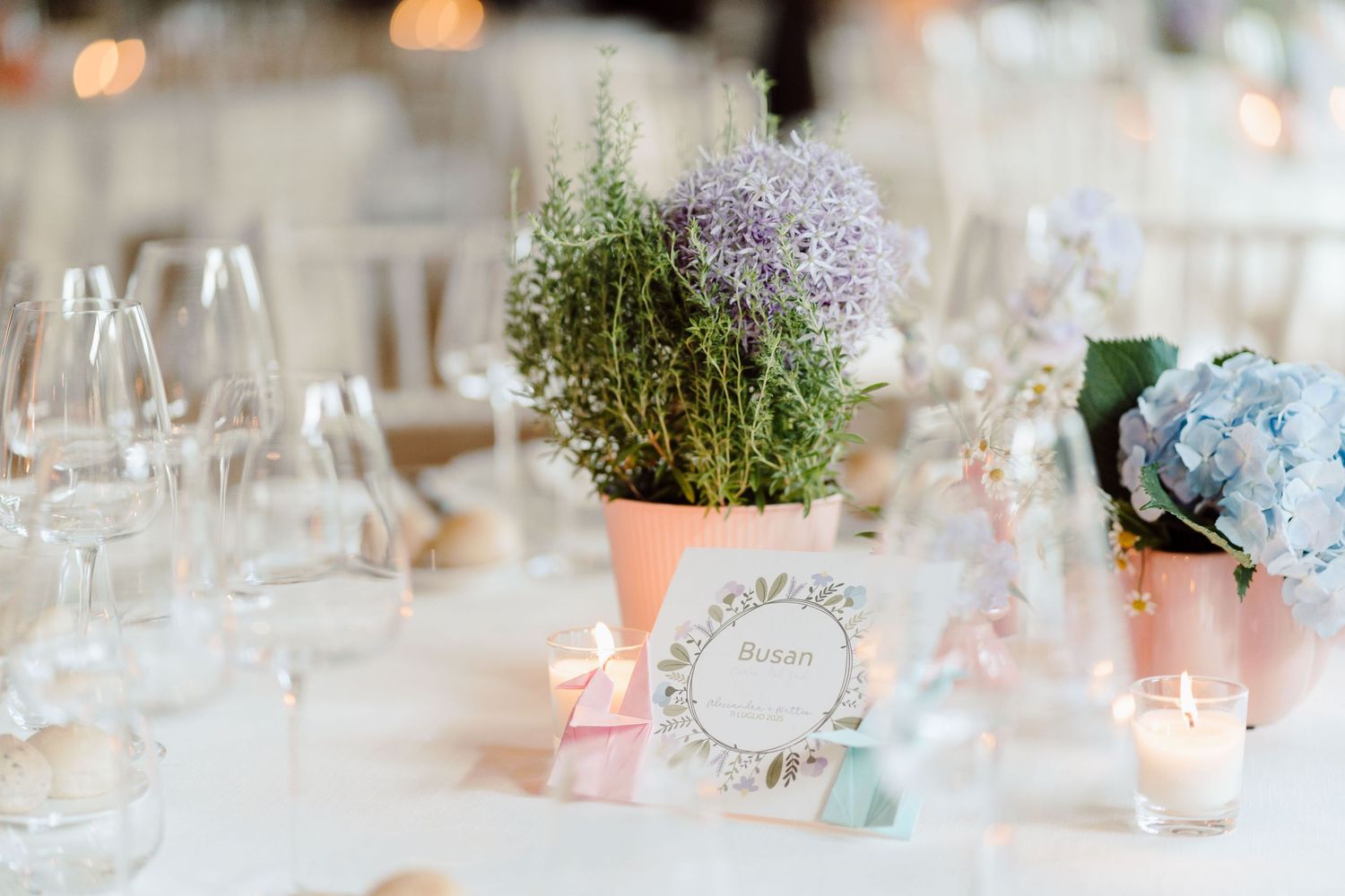 Elegant wedding table centerpiece with potted herbs, blue hydrangeas, candles, and a table name card in soft pastel tones.