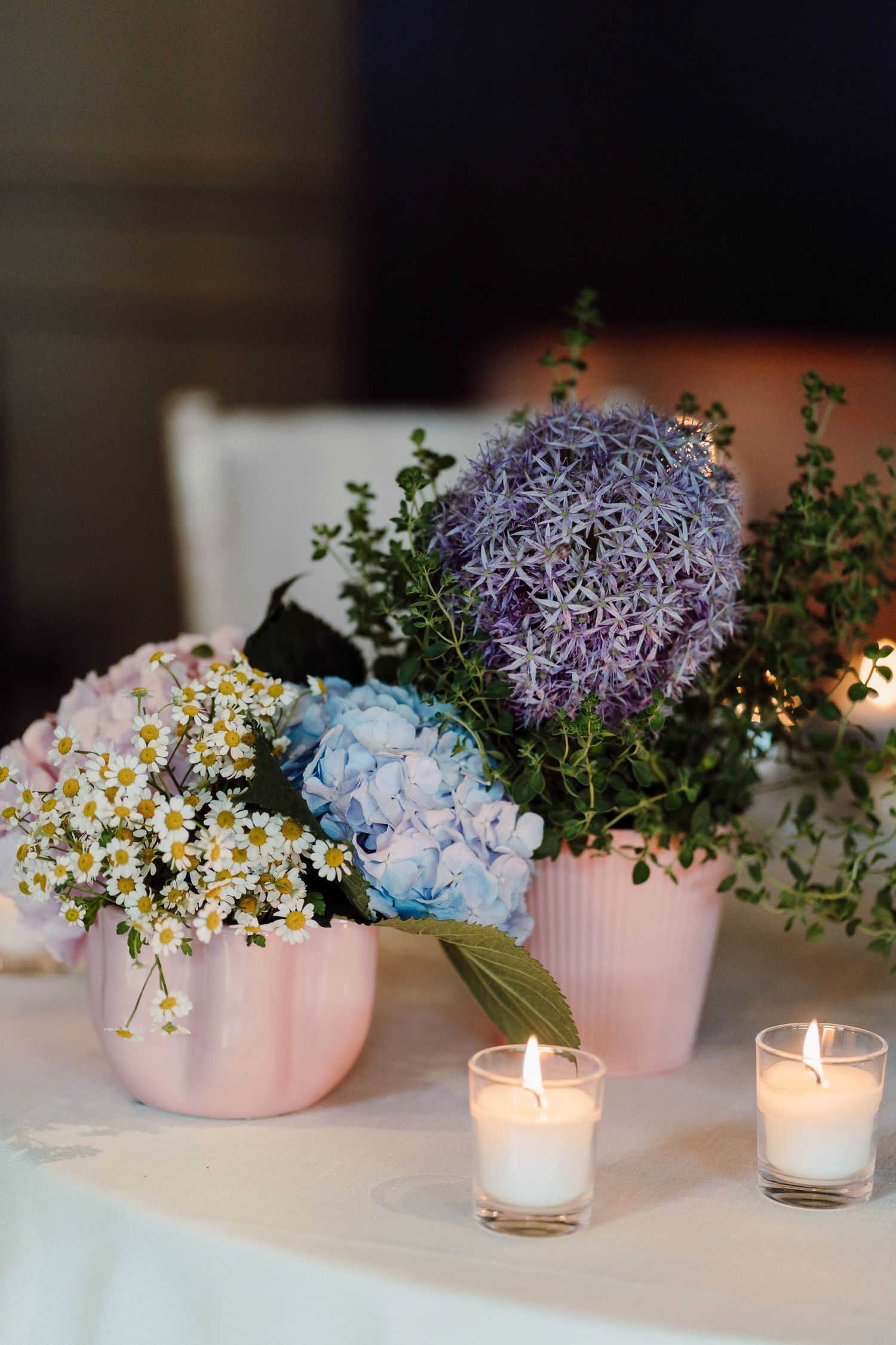Pink pots with blue hydrangeas, purple allium, and white daisies on a white table with lit candles.