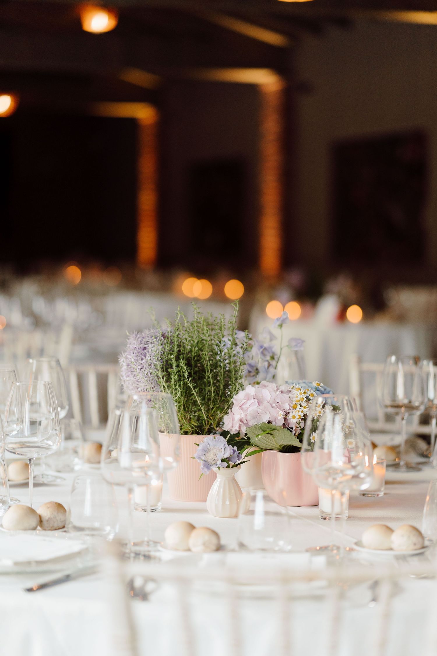 Elegant wedding reception table with pink vases of lavender flowers, crystal glassware, and soft candlelight in a warm venue.