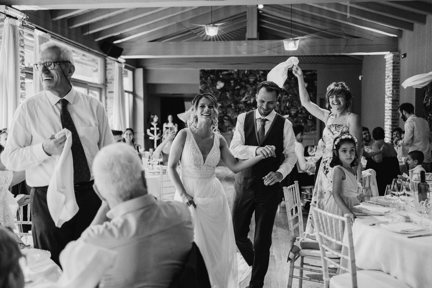 Black and white wedding reception photo of bride and groom dancing joyfully with guests in elegant venue.