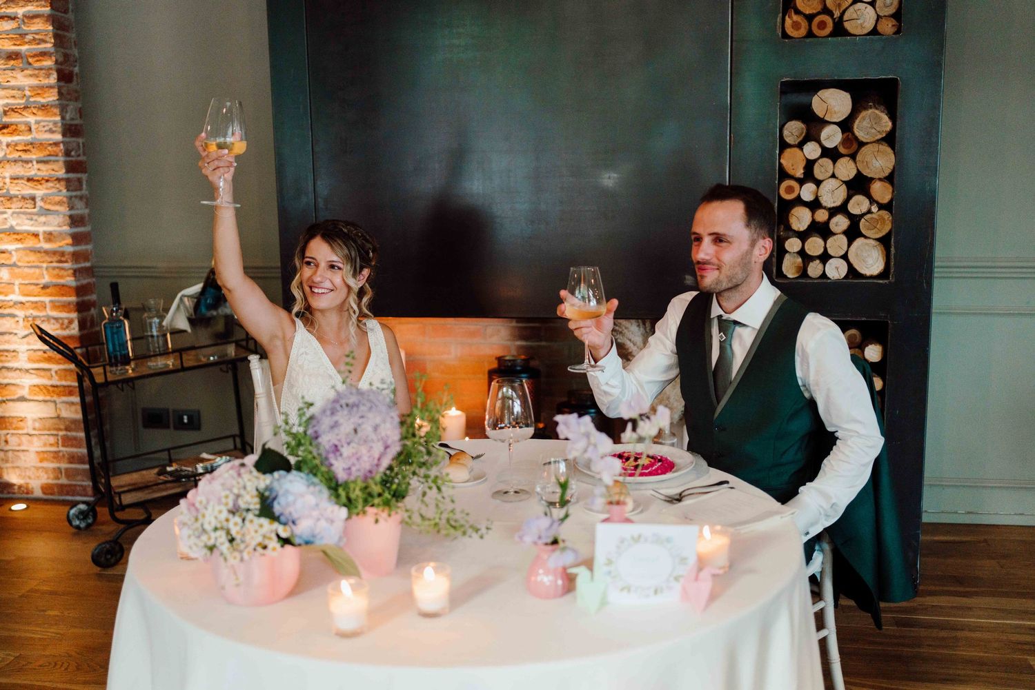 Bride and groom raising glasses in a toast at their sweetheart table decorated with candles and flowers.