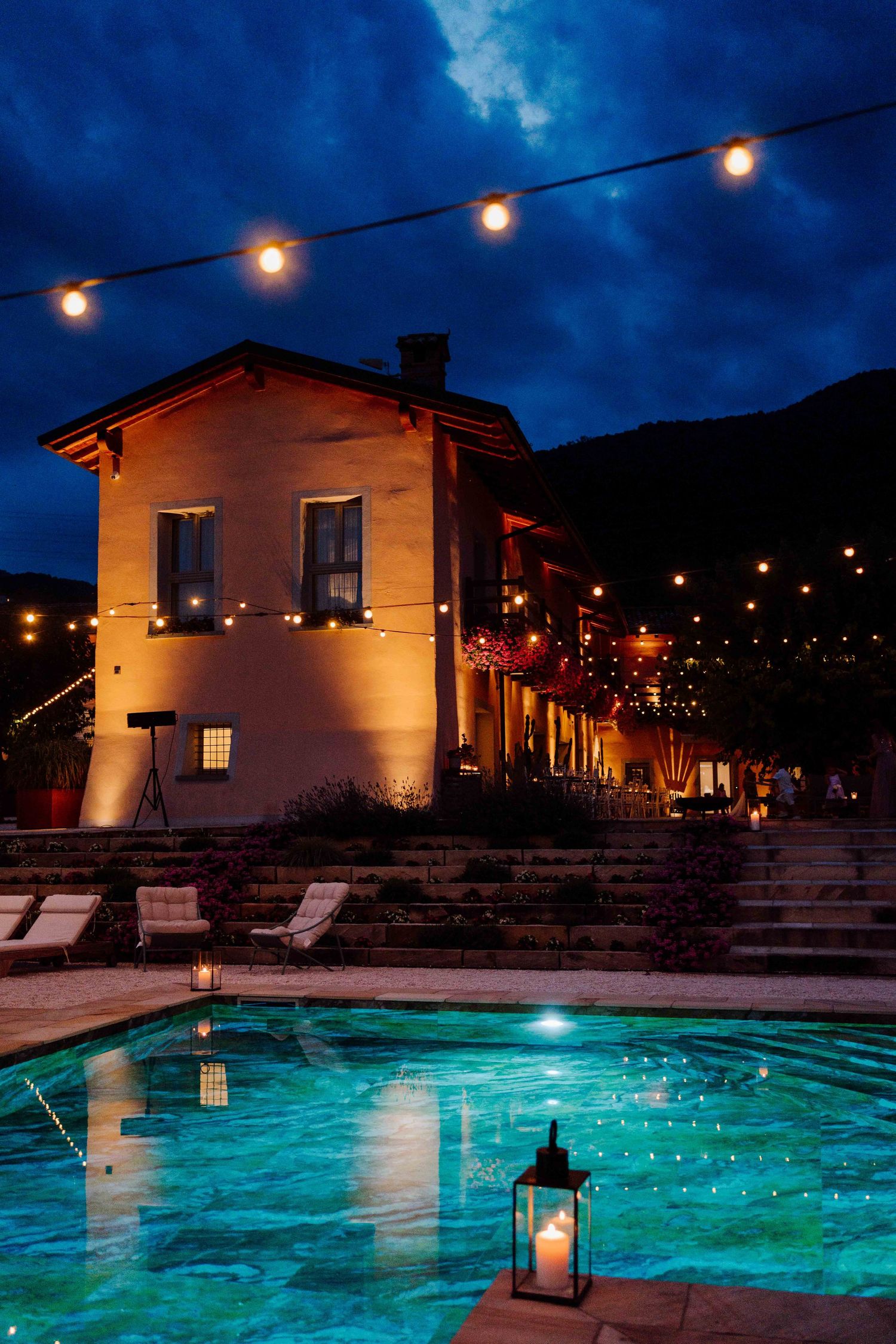Elegant villa with illuminated string lights reflecting in a pool at night, set against a dramatic blue sky.