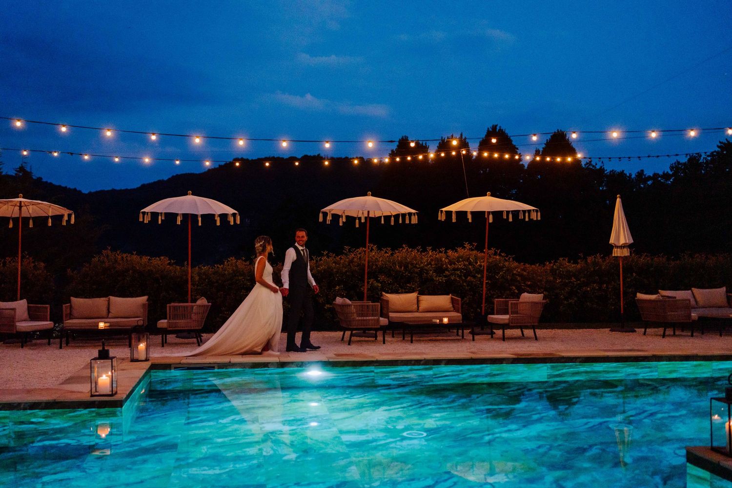 Bride and groom standing by illuminated pool at dusk with string lights and umbrellas at elegant outdoor wedding venue.