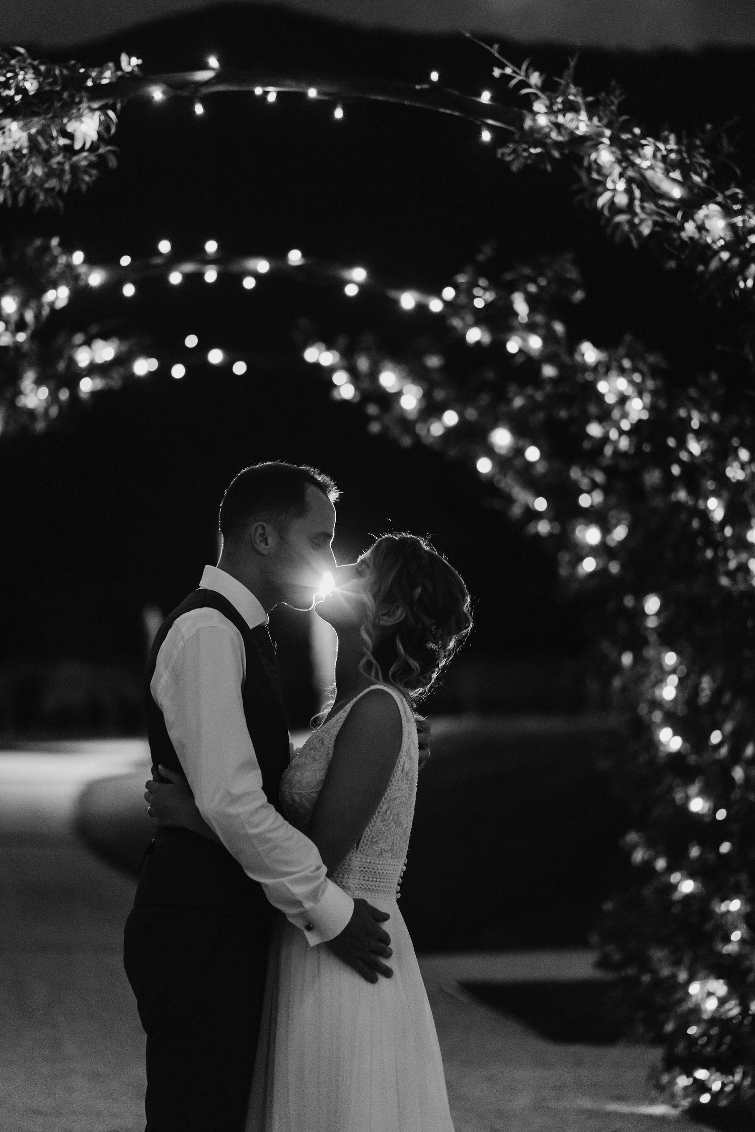 Black and white photo of a couple kissing under a floral arch adorned with twinkling fairy lights at night.