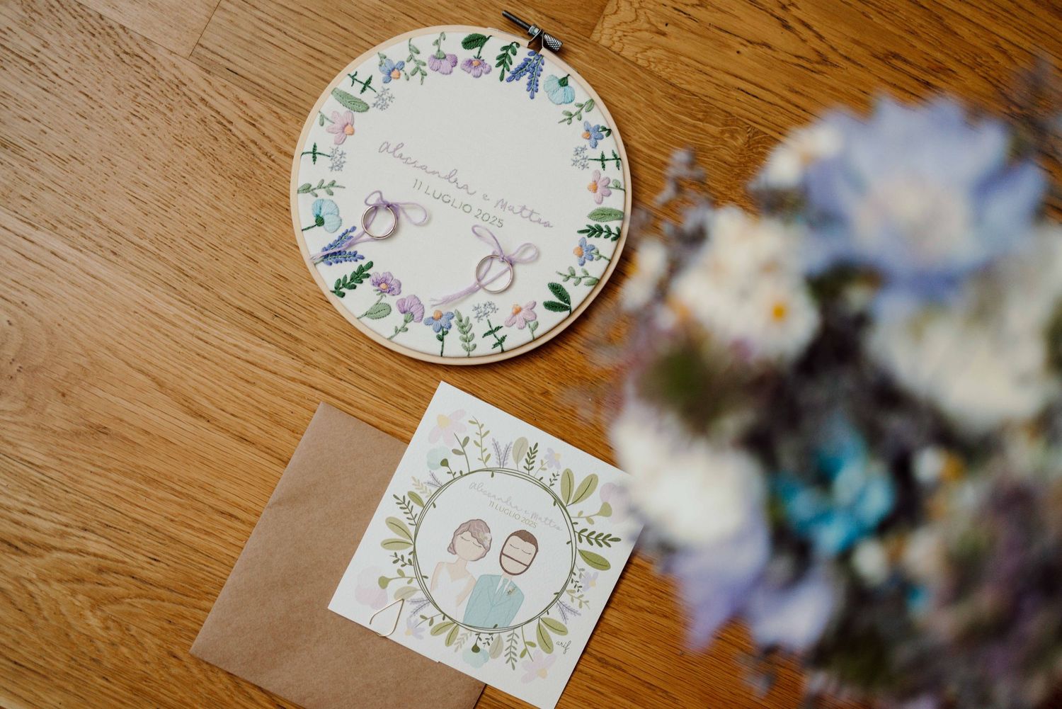 Floral wedding invitations with illustrated couple and rings on wooden floor beside a bouquet of white and blue flowers.