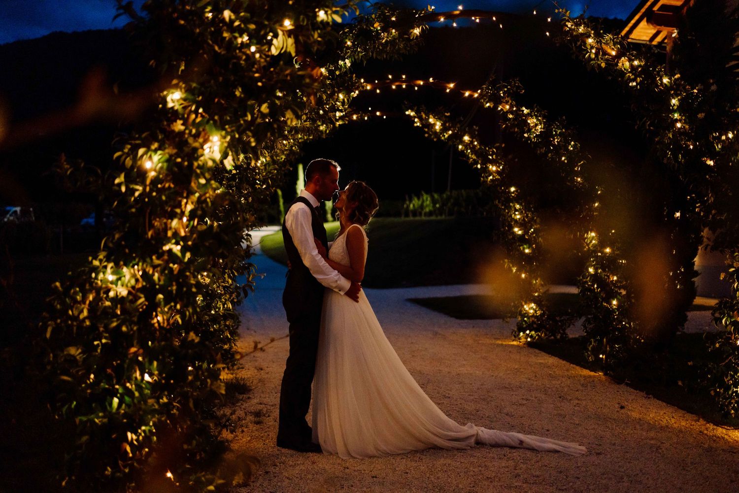 Bride and groom share a romantic moment under a glowing floral arch adorned with fairy lights at night.