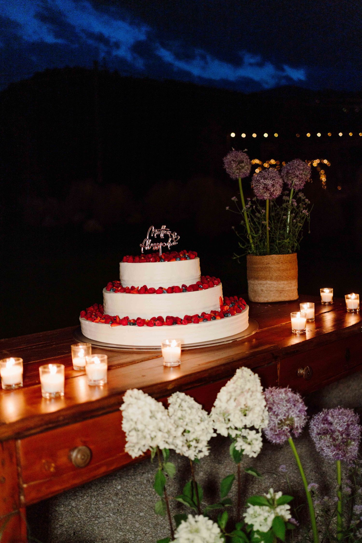 Three-tier white wedding cake with red berries, candlelit table, floral decor, and allium flowers at an outdoor evening reception.