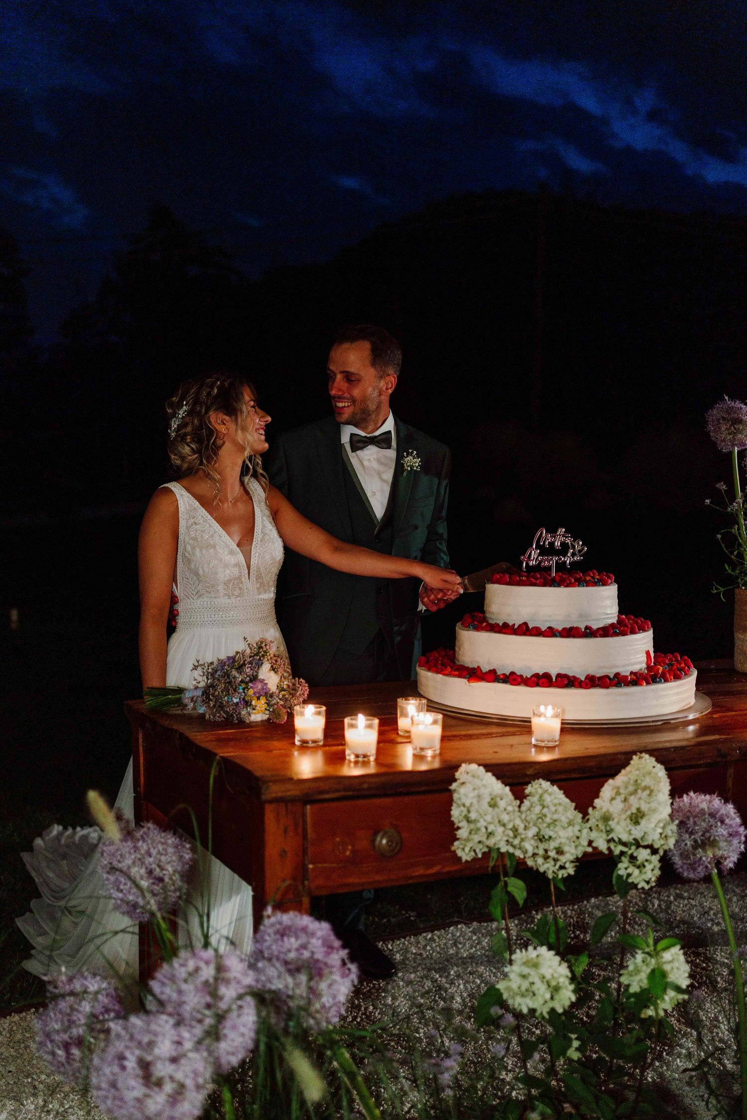 Bride and groom cutting a white wedding cake with red berries by candlelight at an outdoor evening reception.