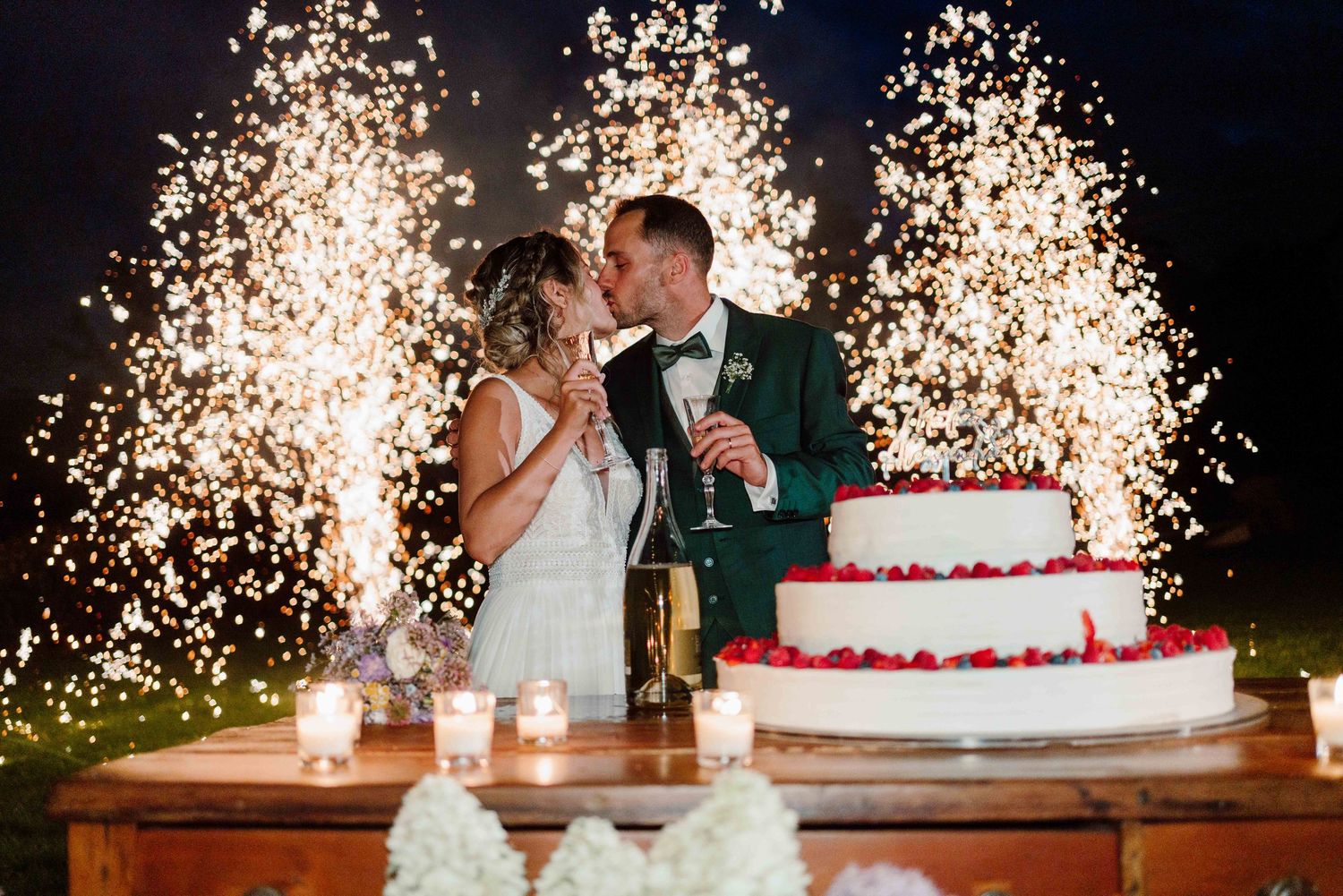 Bride and groom kissing at wedding cake table with dramatic fountain sparklers at elegant night reception.