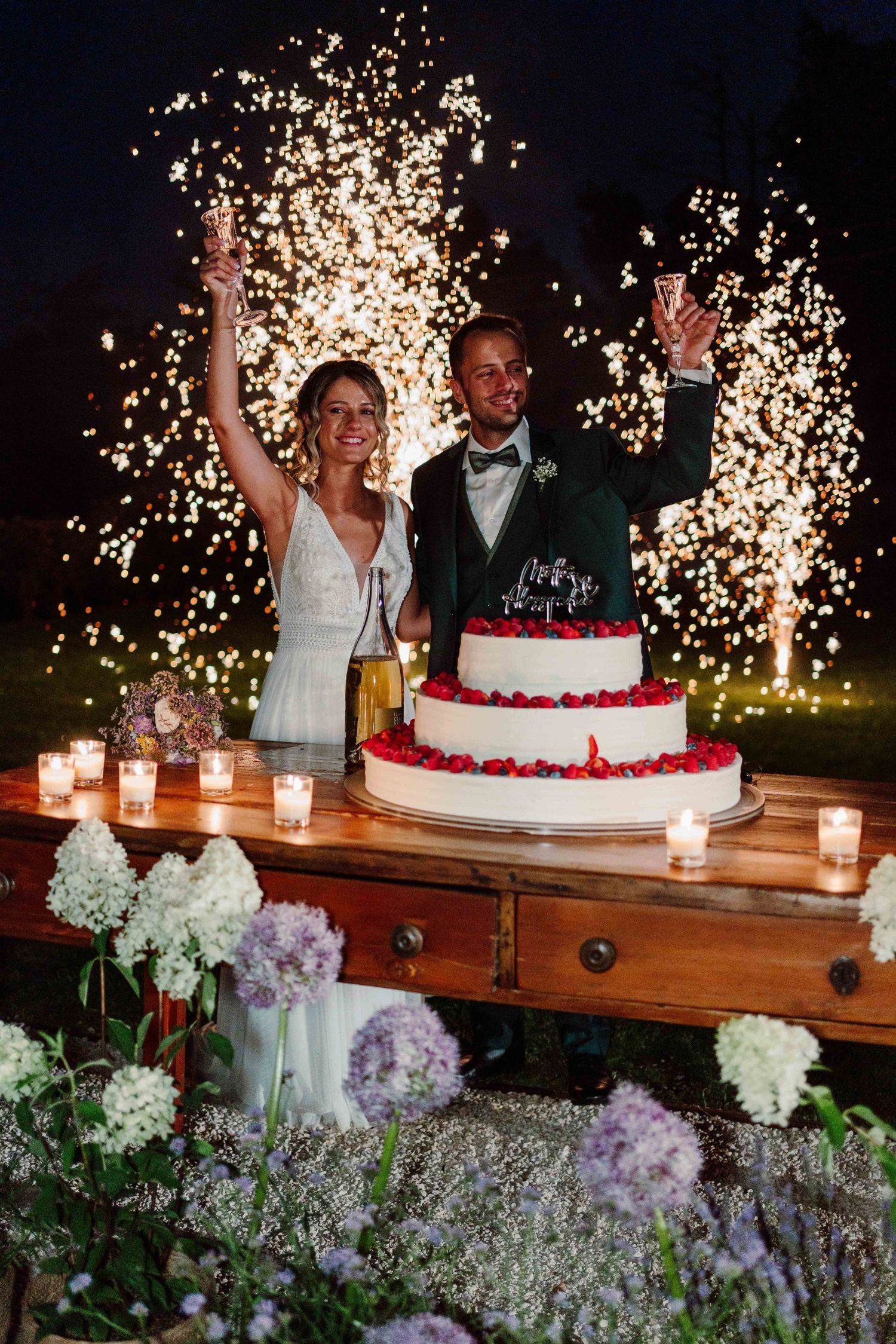 Bride and groom celebrating wedding cake cutting with sparkling fireworks and candles at elegant outdoor night reception.