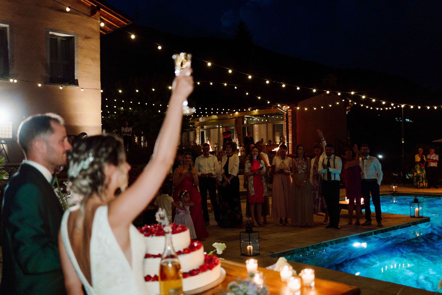 Bride raises a glass in toast next to groom and wedding cake at elegant outdoor night reception with string lights and pool.