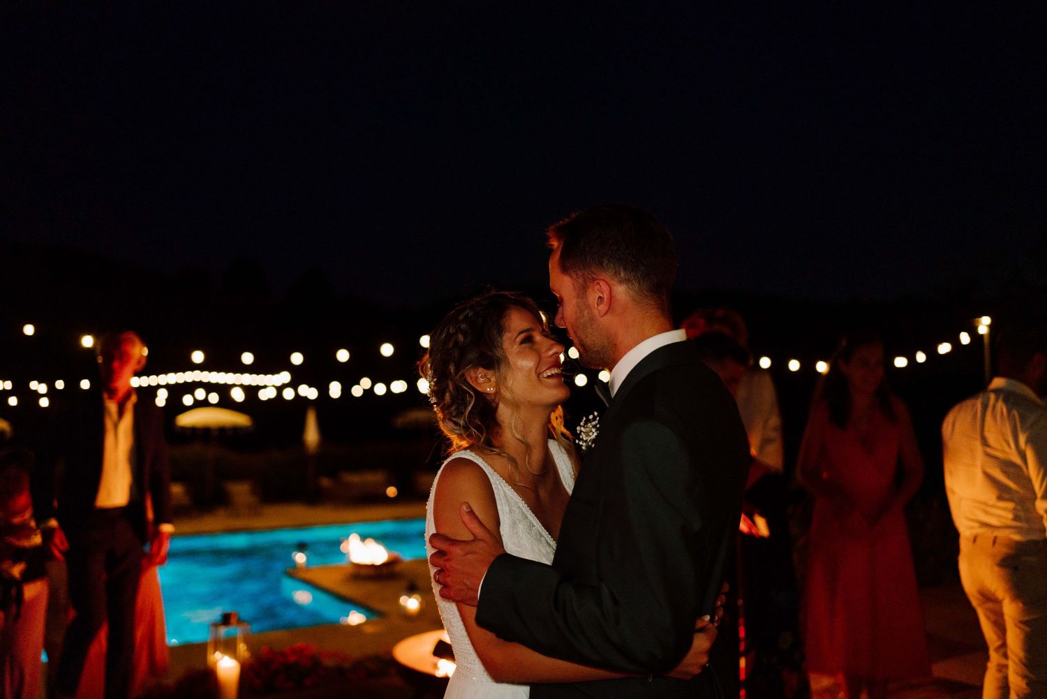 Couple sharing a romantic kiss at an outdoor evening wedding reception with string lights and pool in background.