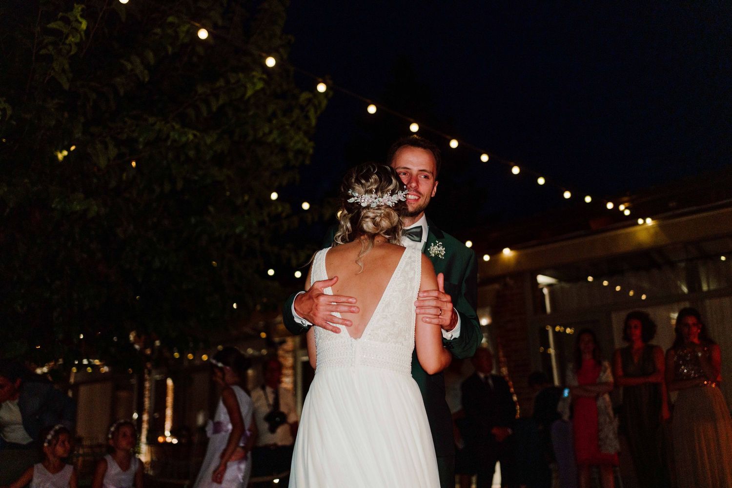 Bride and groom share their first dance outdoors at night under warm string lights at a romantic wedding reception.
