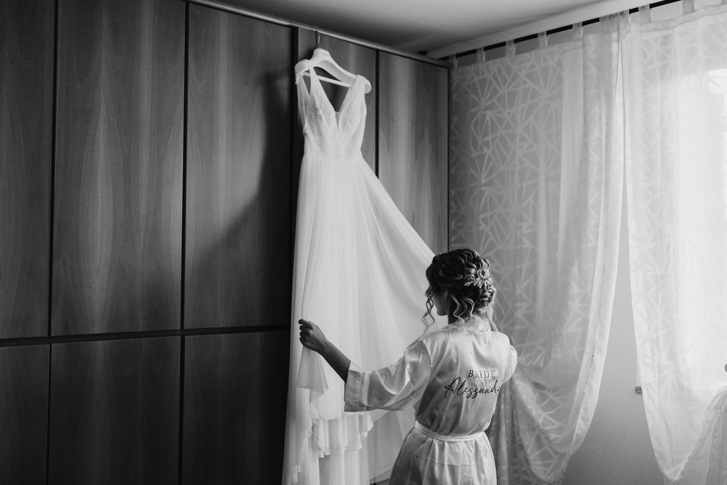 Bride in robe admiring her white wedding dress hanging on a wooden wardrobe in a softly lit room.