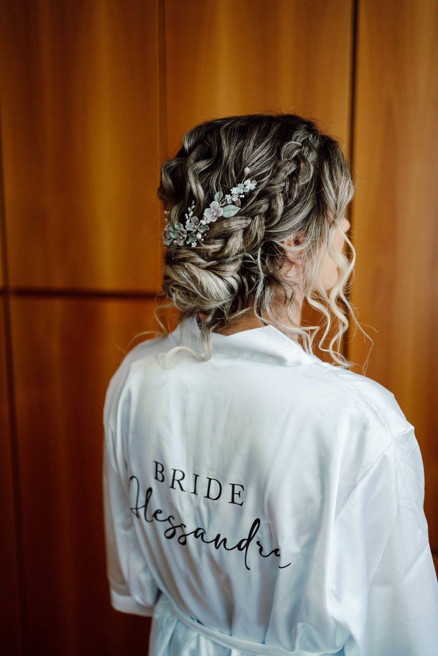 Bride Alessandra wearing a white satin robe with an elegant braided updo adorned with floral hair accessories.