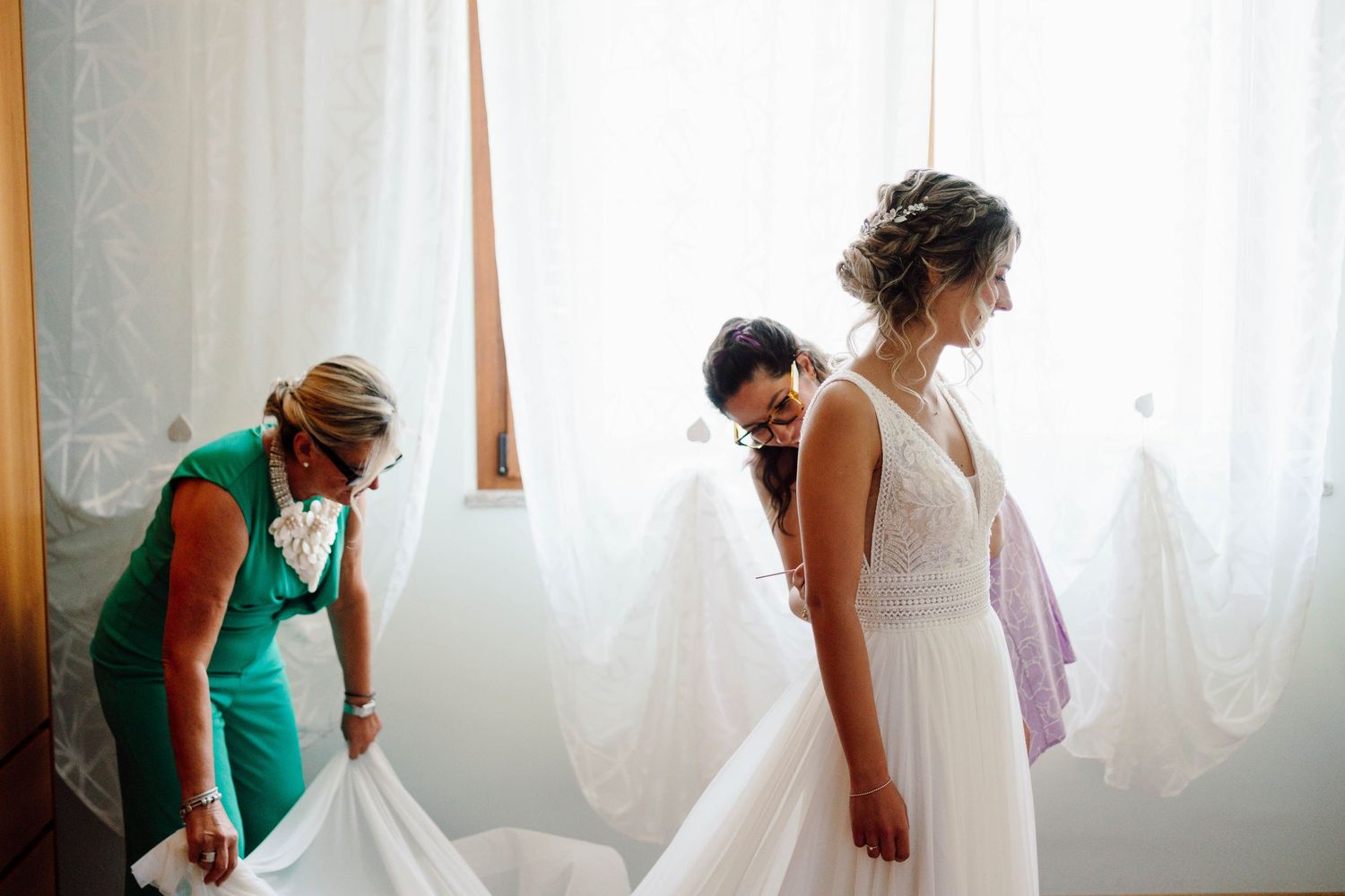 Bride in white gown being helped by two women in elegant room with sheer curtains before wedding ceremony.