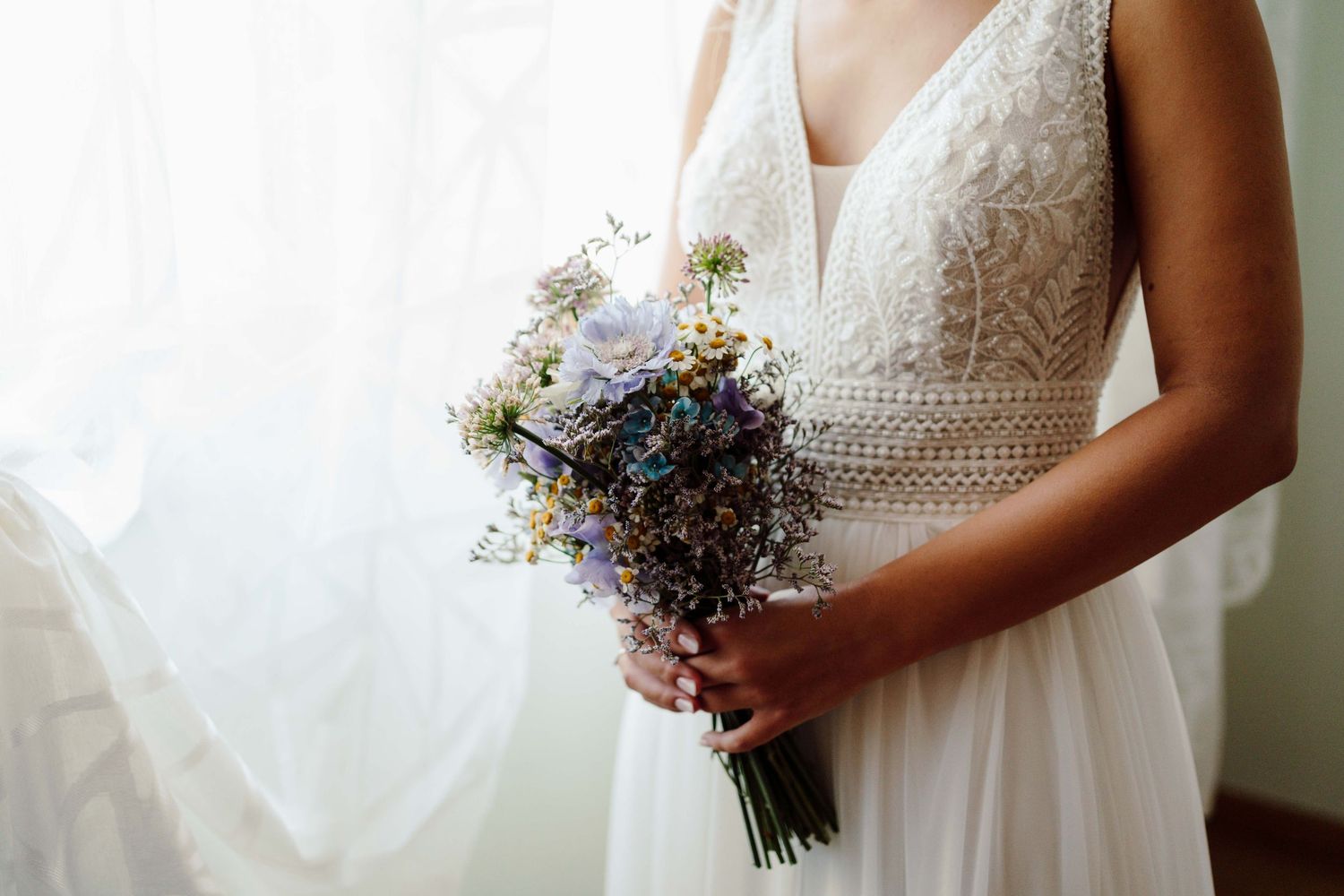 Bride in white lace wedding dress holding a delicate bouquet of blue and white wildflowers near a bright window.