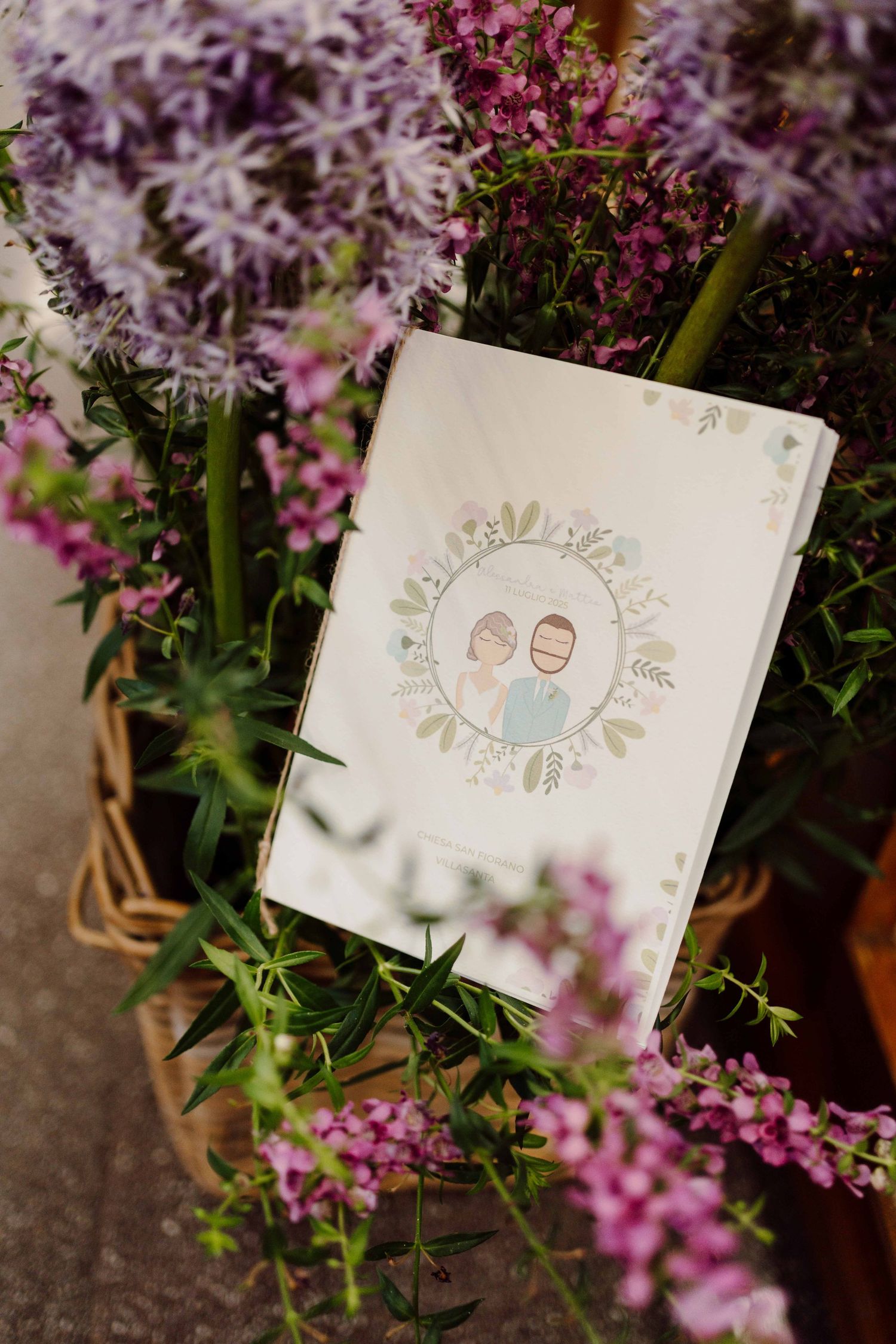 Wedding card with illustrated couple portrait surrounded by floral wreath, nestled among purple and pink flowers in a wicker basket.