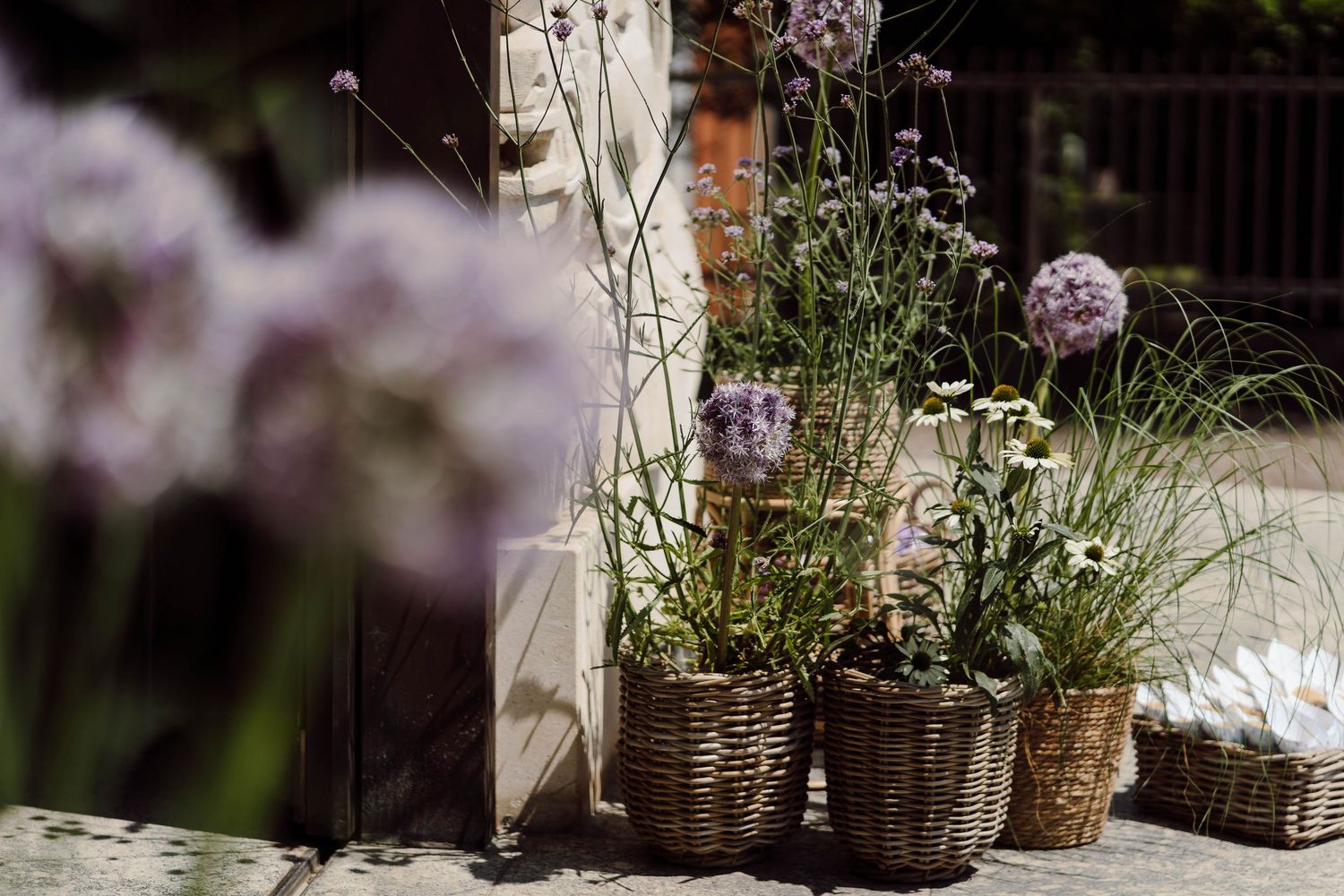 Wicker baskets filled with allium and wildflowers arranged on a sunny patio beside a rustic wooden post.