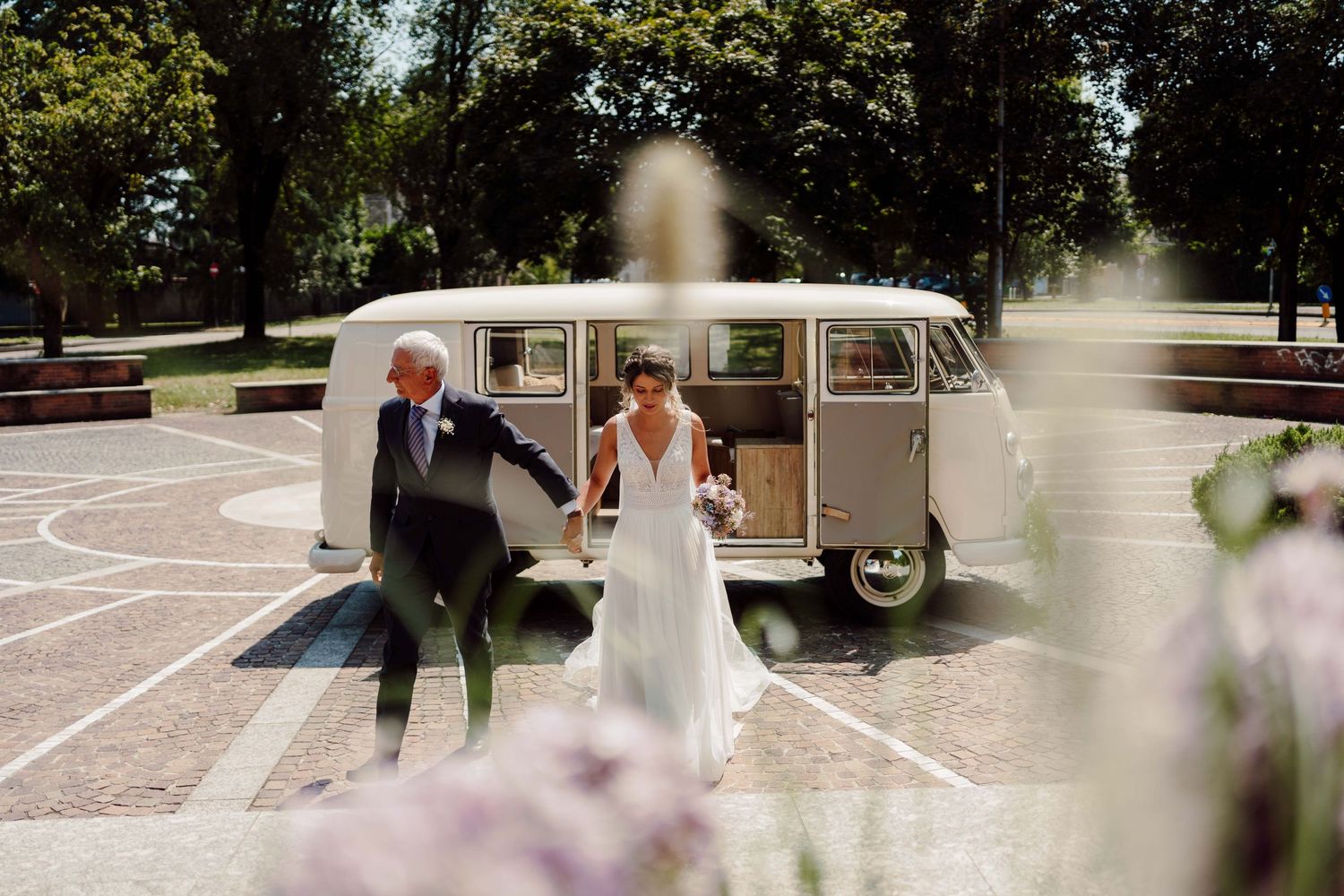 Bride and groom walking hand in hand in front of a vintage VW camper van at an outdoor wedding venue.