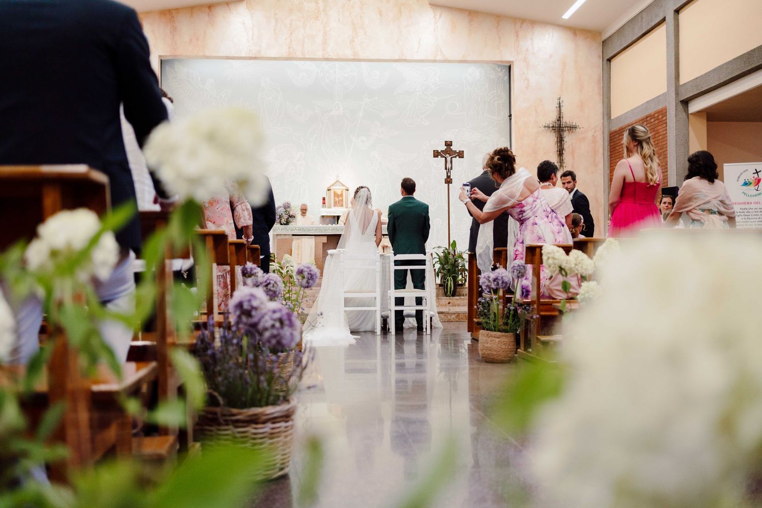 Elegant church wedding ceremony with floral decorations, couple at altar, guests seated in pews, cross visible on wall.