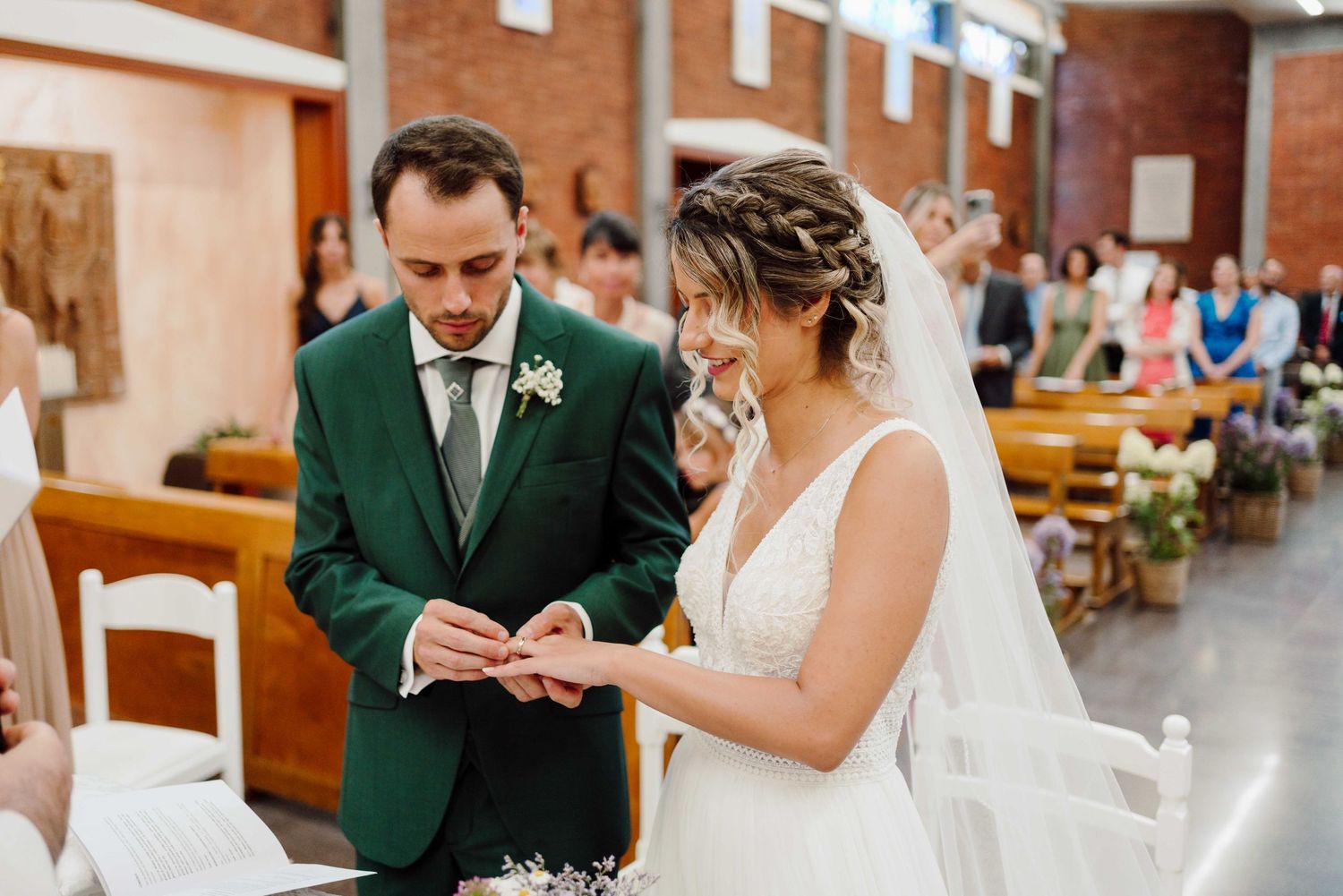 Groom in green suit places wedding ring on bride's finger during church ceremony.
