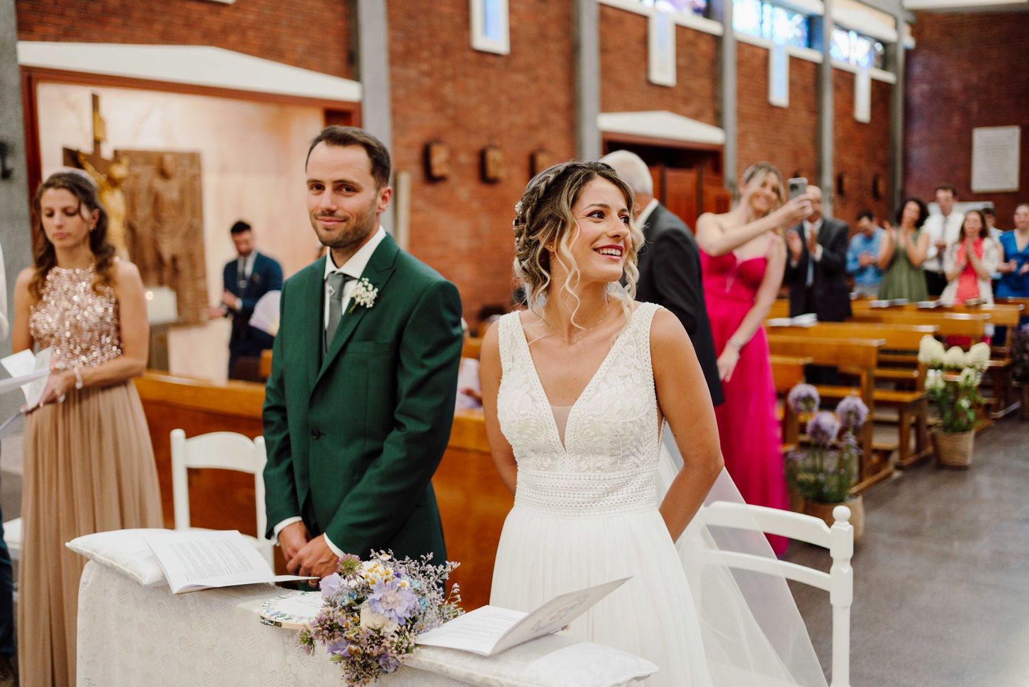 Smiling bride and groom stand at the altar during a church wedding ceremony with guests seated in pews behind them.