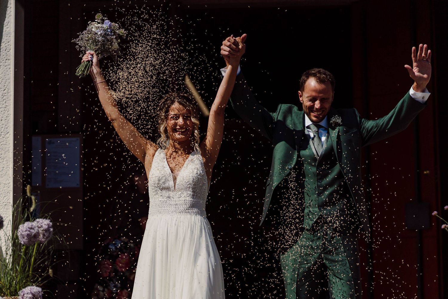 Joyful newlyweds celebrate outside with confetti, bride in white gown holding bouquet, groom in green suit raising hands.