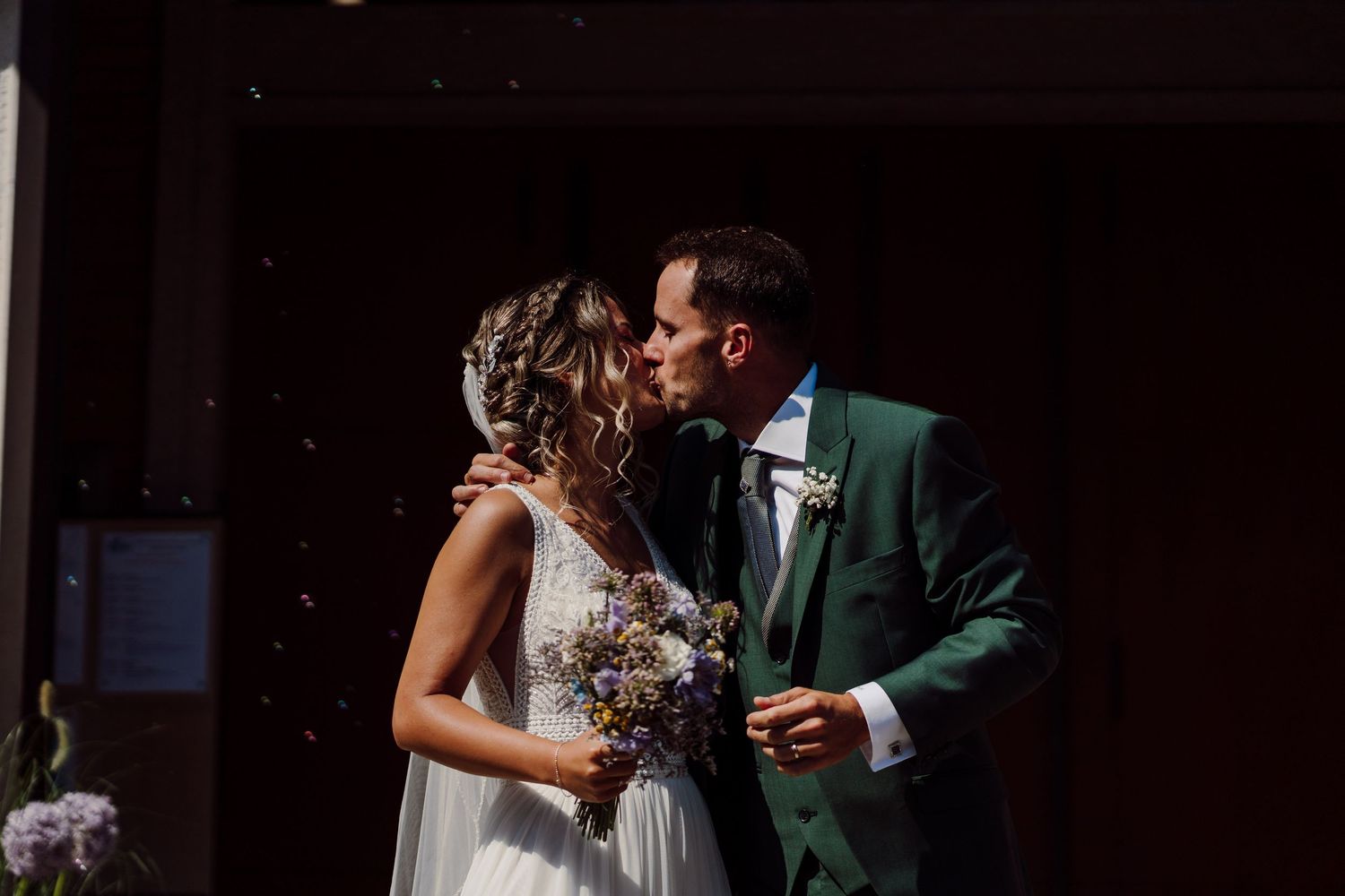 Bride and groom share a first kiss outdoors, bride holding wildflower bouquet, groom in green suit.