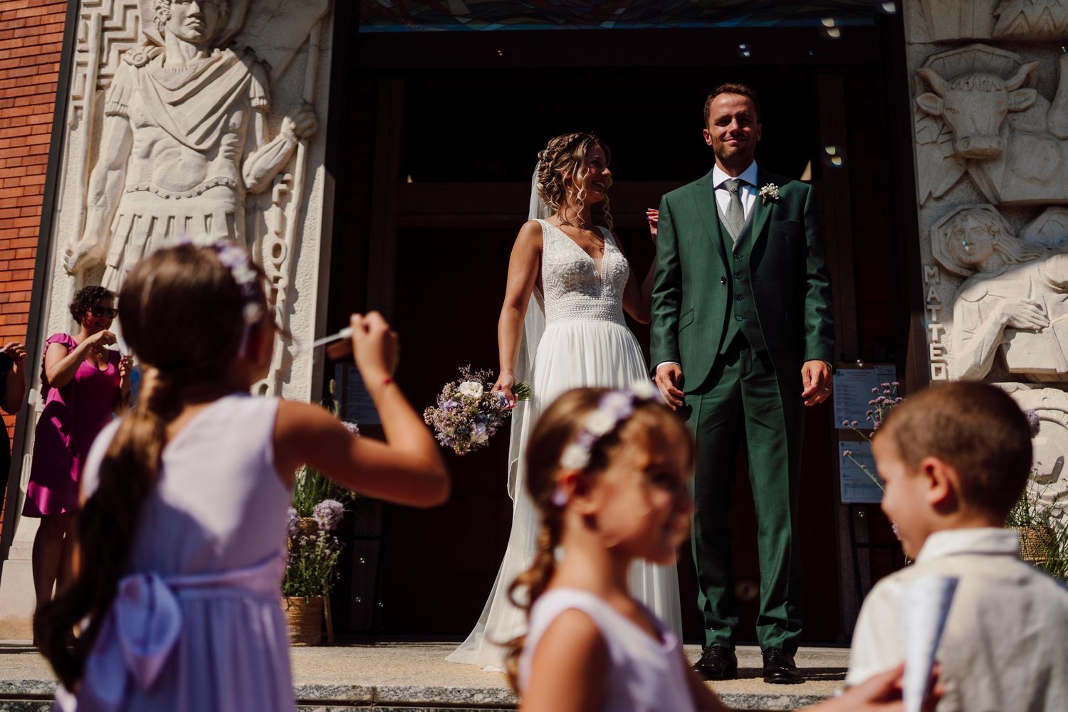 Bride and groom exit church greeted by children with wands at sunny outdoor wedding ceremony.
