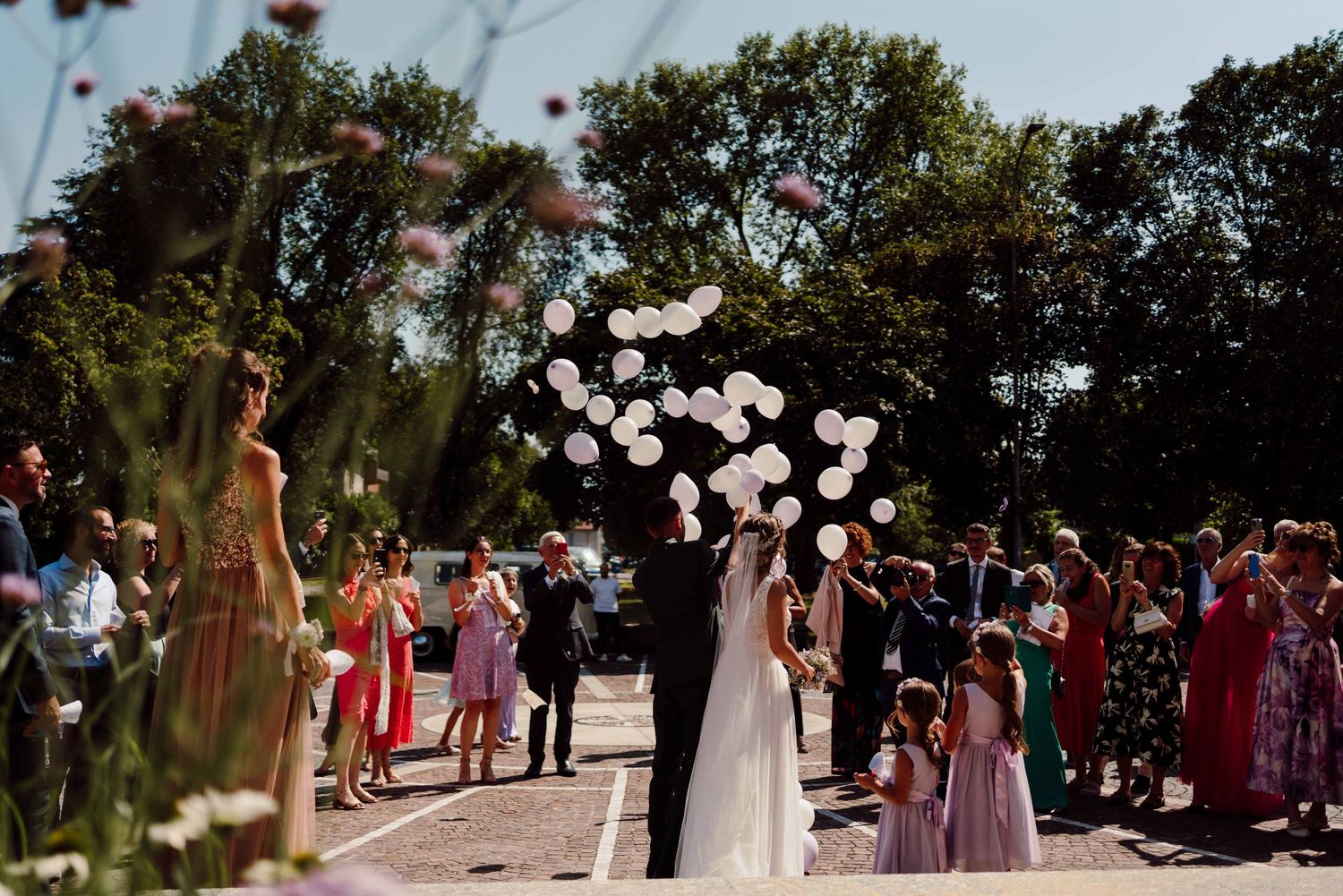 Bride and groom release white heart balloons surrounded by wedding guests outdoors on a sunny day.