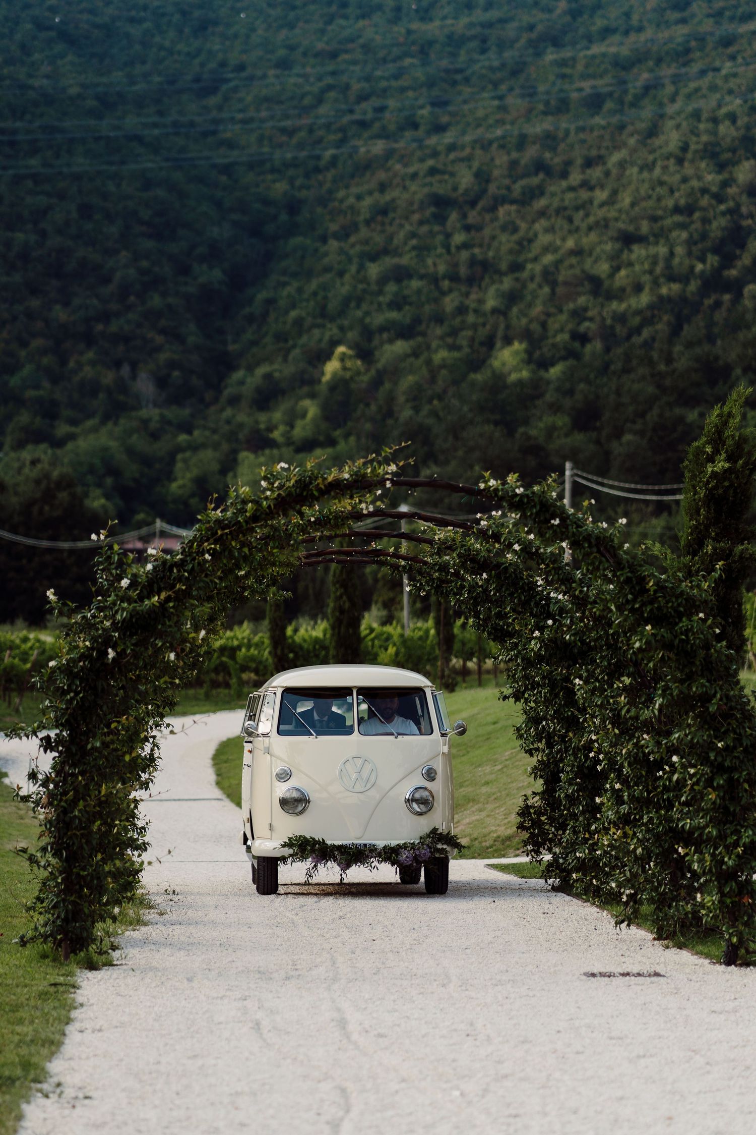 Vintage white VW camper van drives through a floral arch on a gravel path with lush green hills in background.