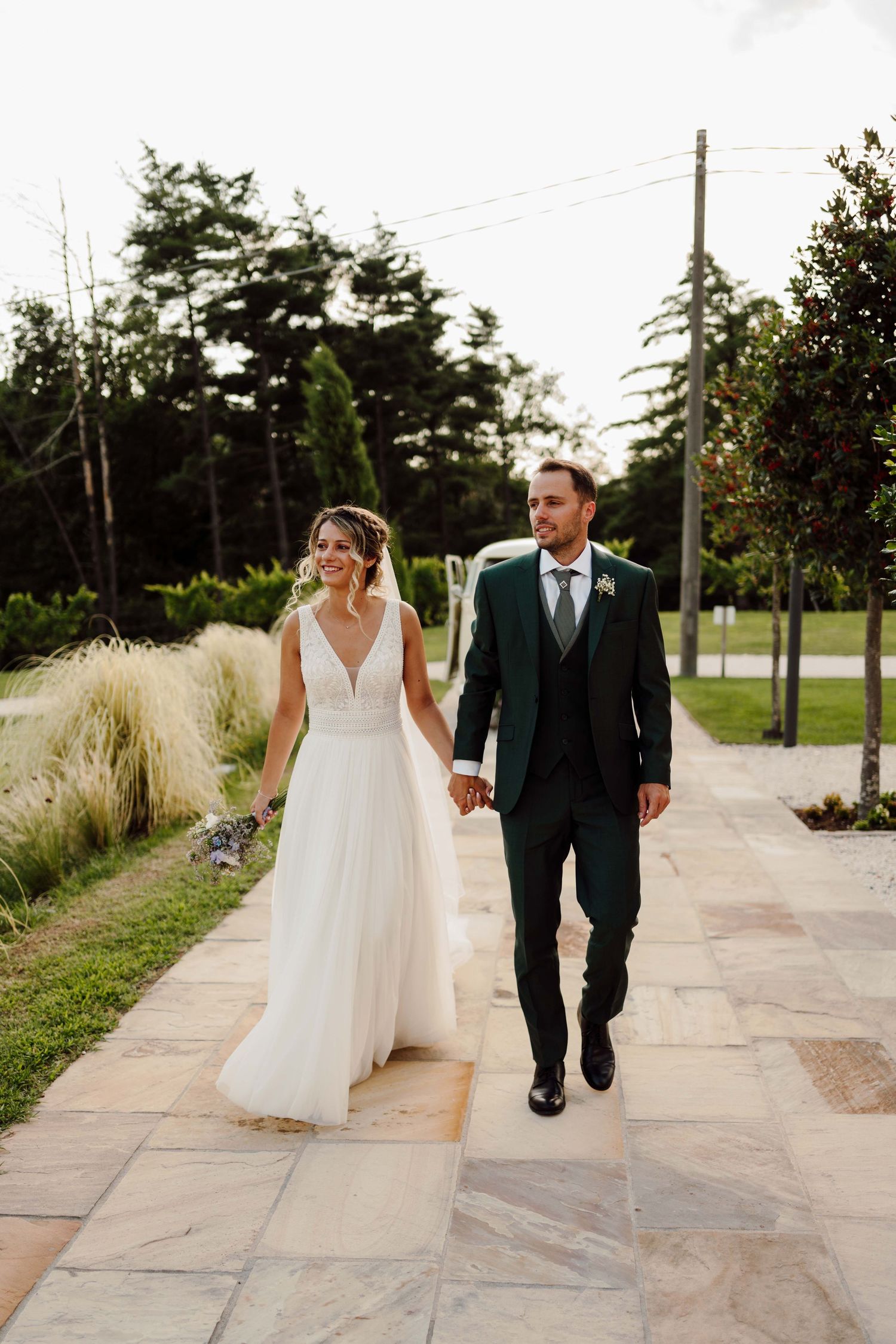 A smiling bride in a white gown and groom in a dark green suit hold hands while walking along a stone path outdoors.