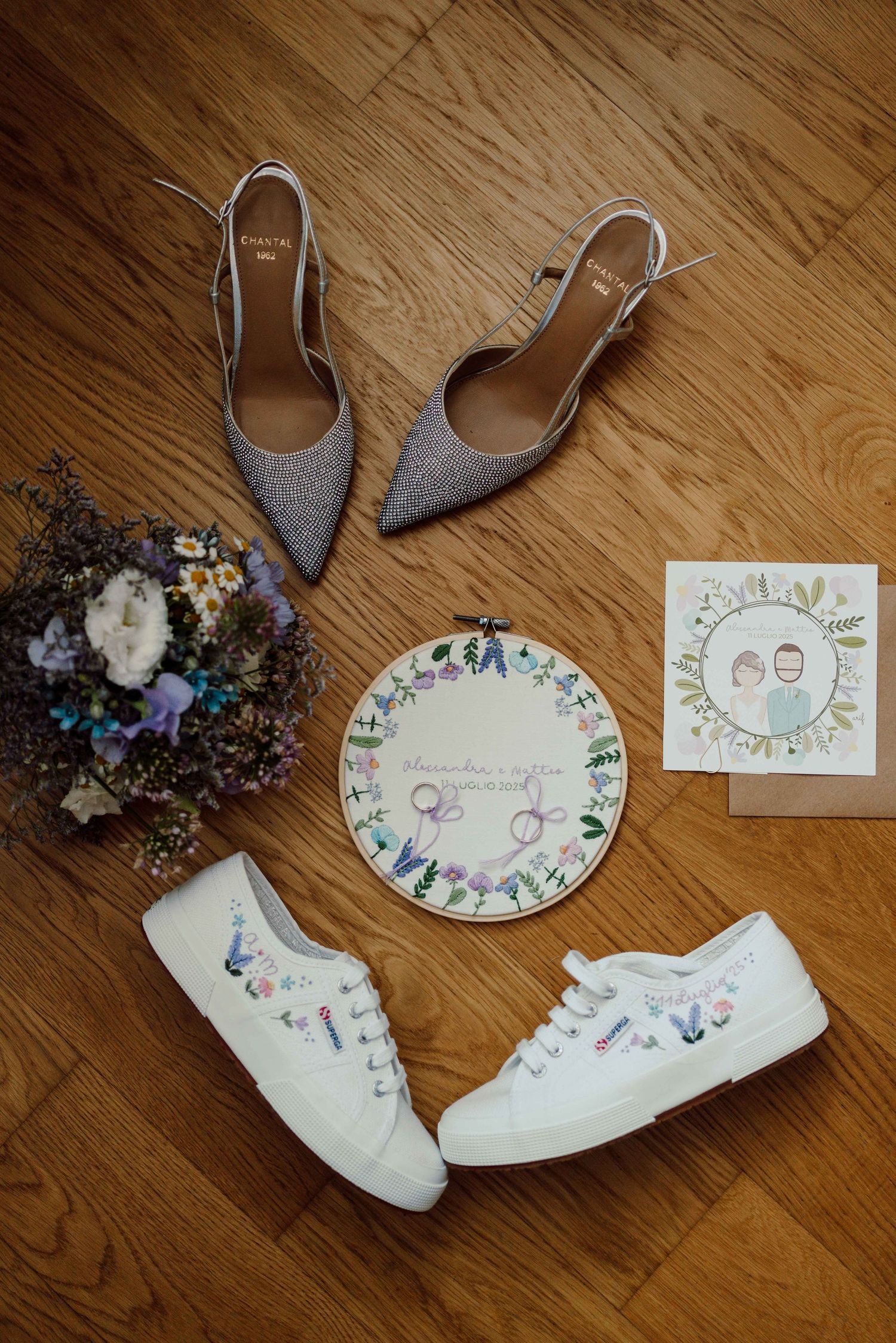Flat lay of wedding shoes, white sneakers, floral bouquet, embroidery hoop, and illustrated cards on wood floor.