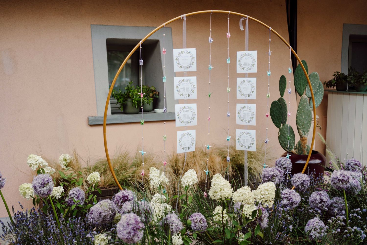 Gold hoop wedding arch with lace details surrounded by purple allium and white hydrangea flowers near a cactus.