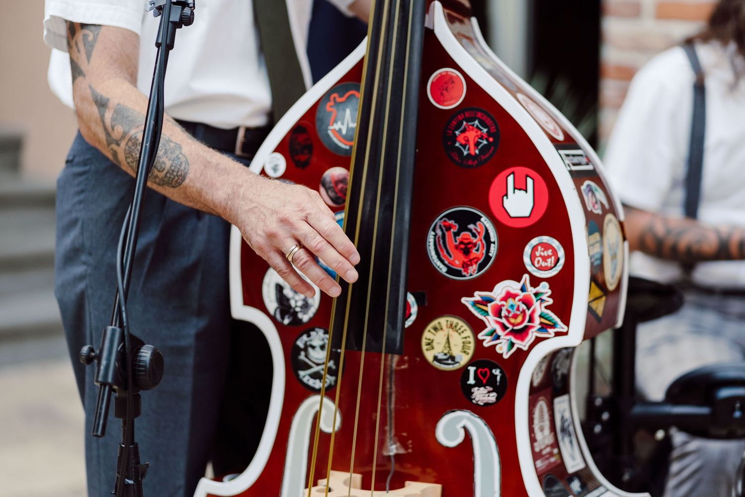 Tattooed musician playing a red double bass decorated with colorful pins and patches at an outdoor event.