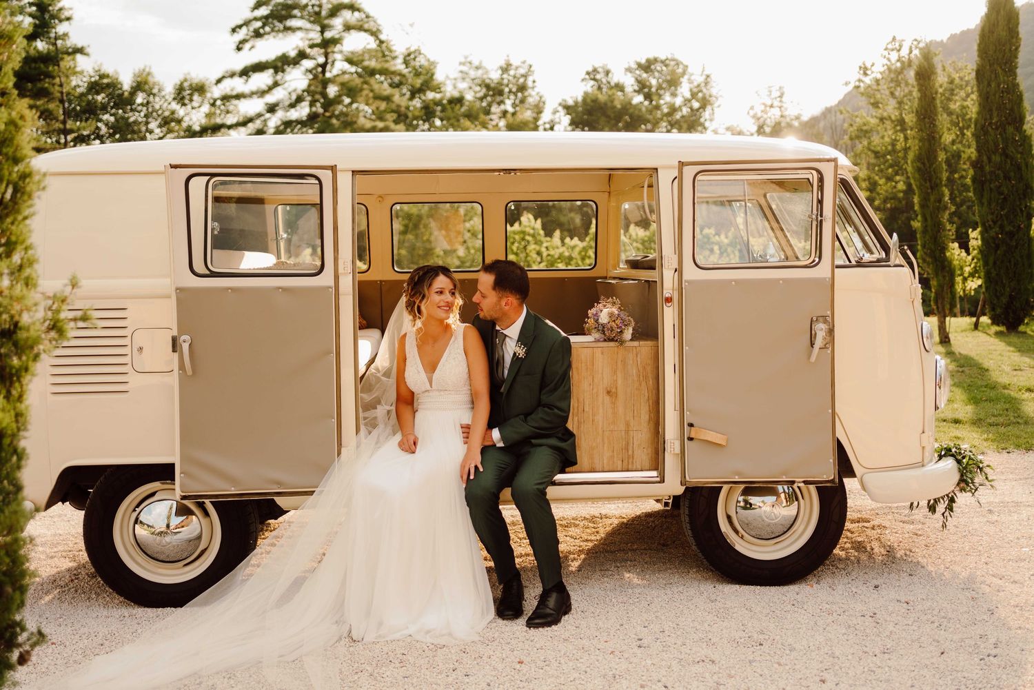 Bride and groom share romantic moment seated outside vintage VW camper van at outdoor wedding venue.