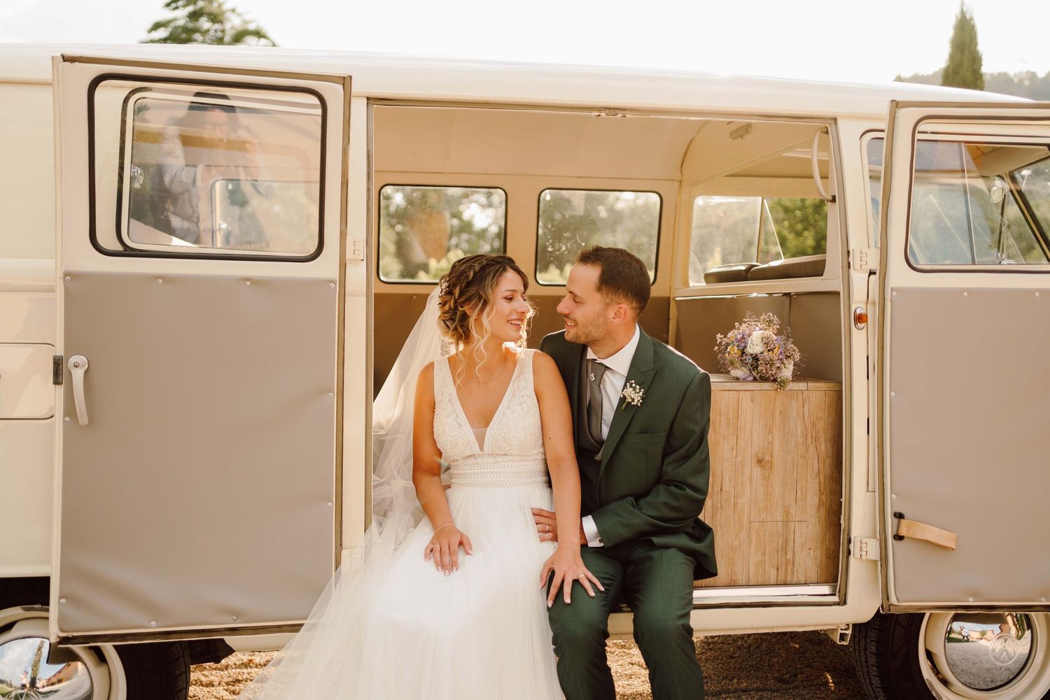 Bride and groom sitting together smiling in front of a vintage cream VW camper van on their wedding day.