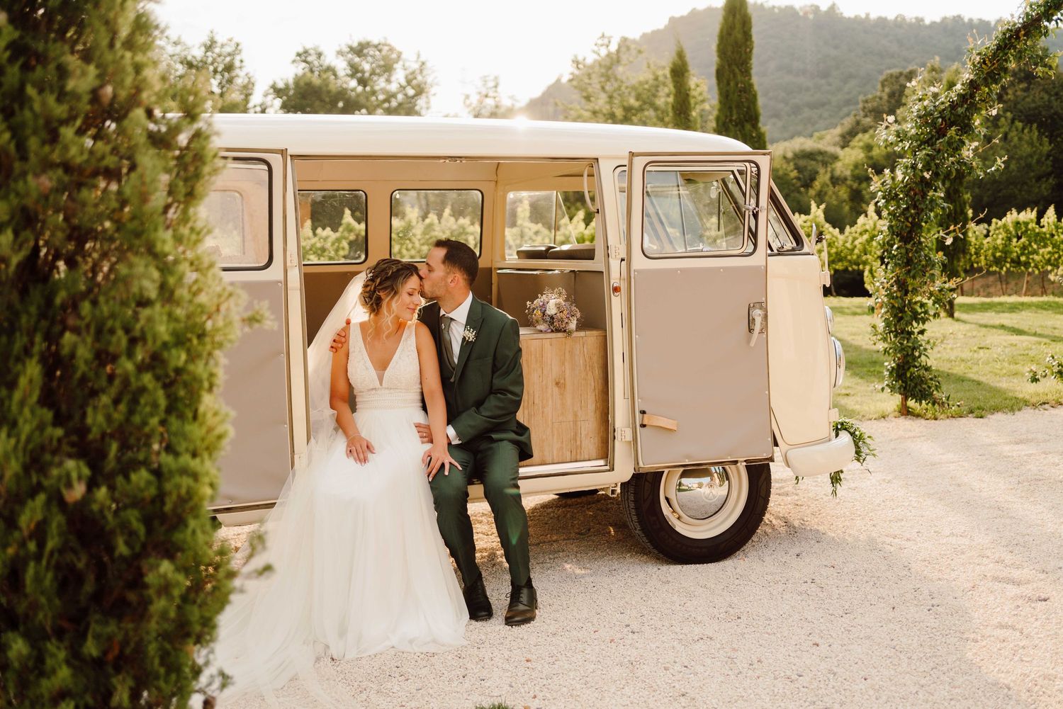 Bride and groom share a kiss beside a vintage cream VW camper van at an outdoor vineyard wedding venue.