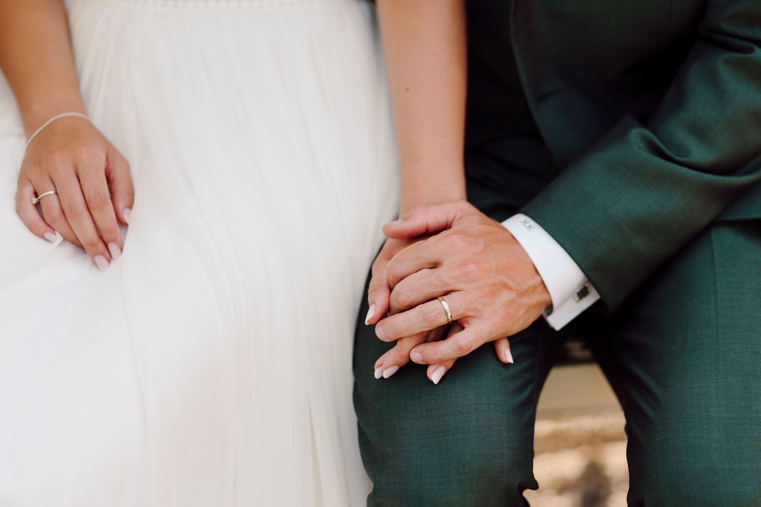 Bride and groom holding hands showing wedding rings, wearing white dress and dark green suit.