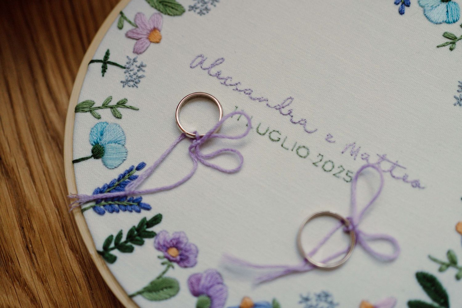Embroidered hoop with floral design, wedding rings tied with purple ribbon, reading Alexandra e Matteo 11 Luglio 2025.