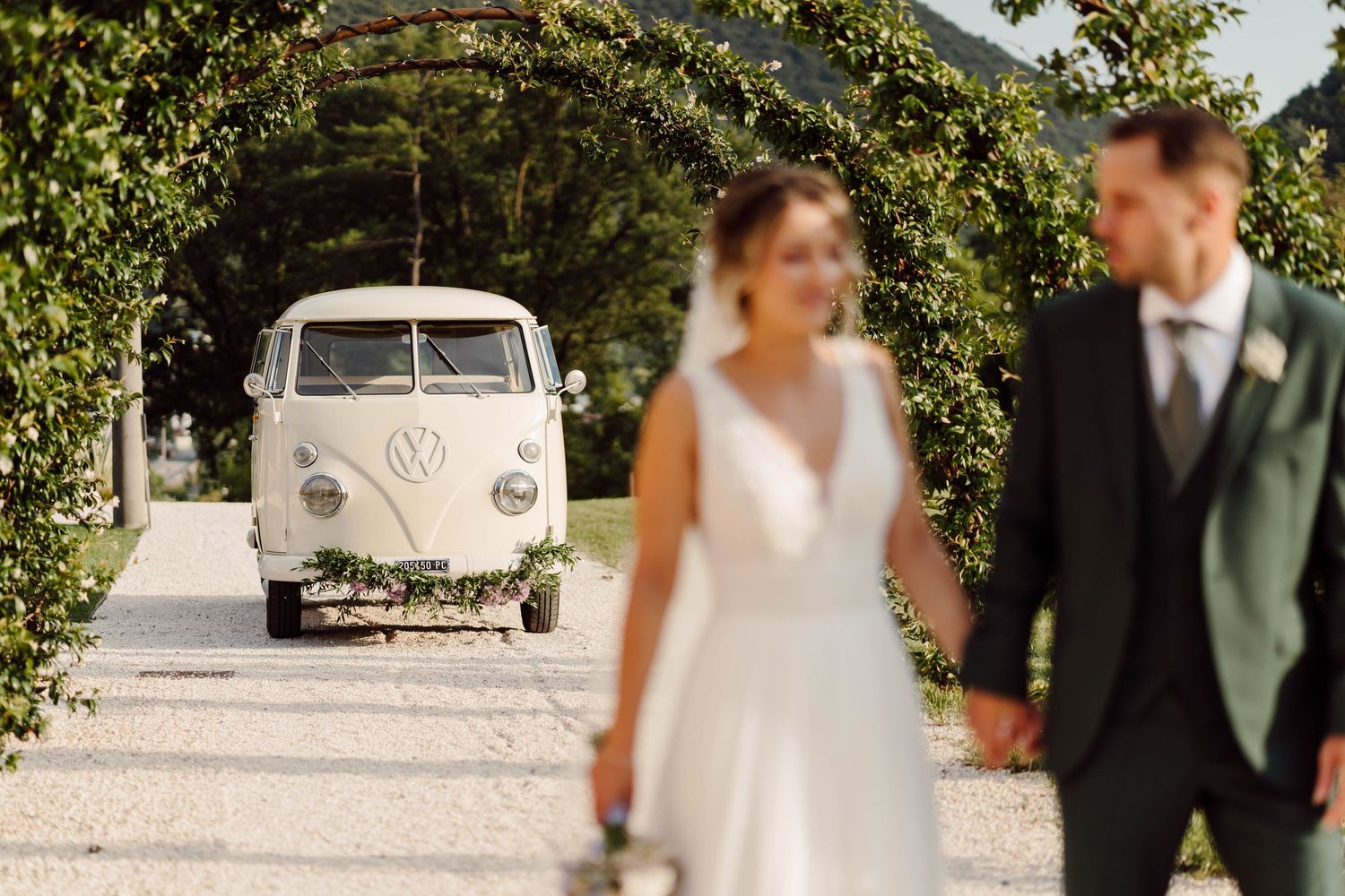 Bride and groom hold hands with a vintage VW camper van on their wedding day in a lush green garden setting.